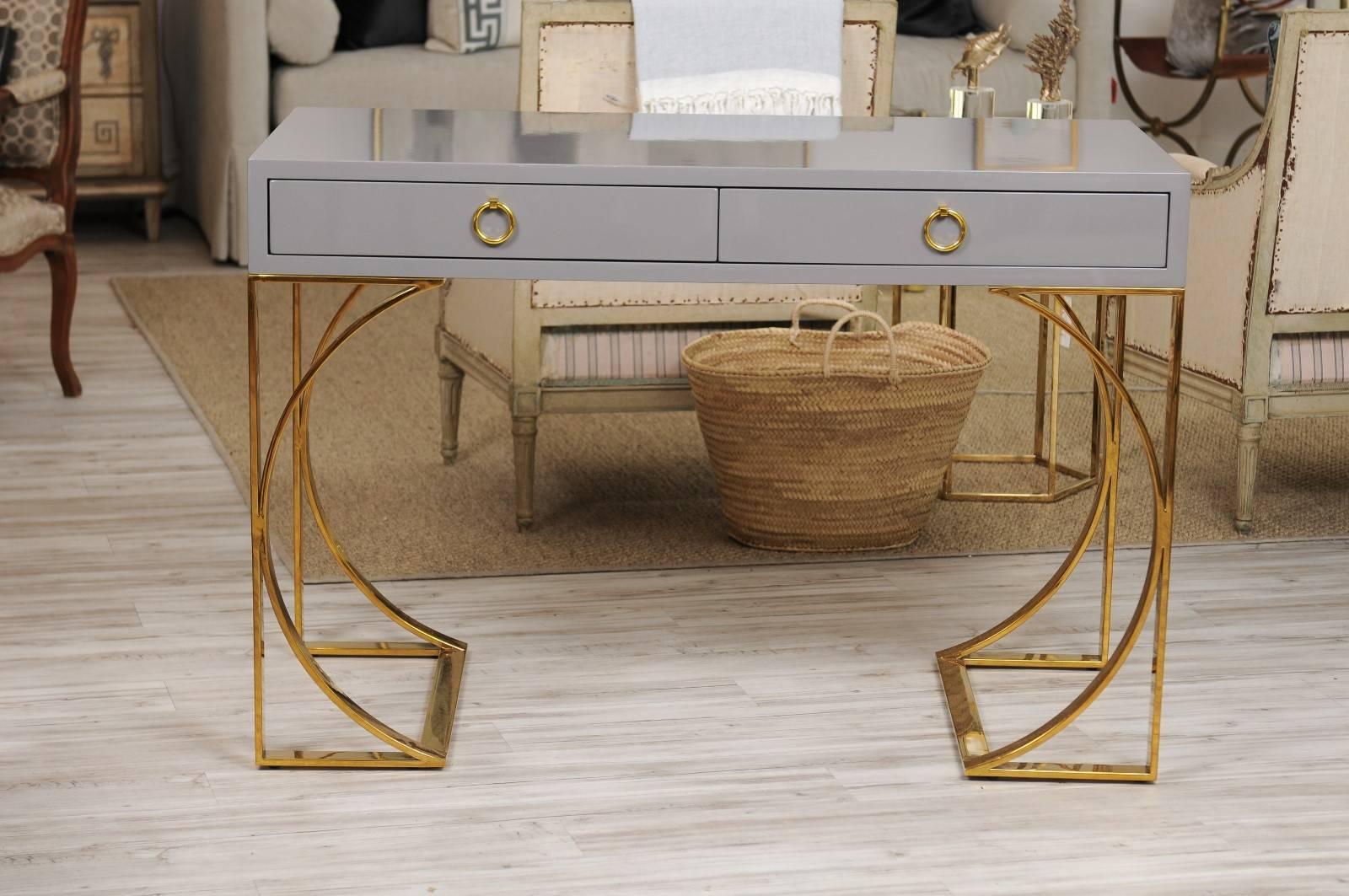 A sophisticated grey lacquer two-drawer desk with gleaming brass arched base. The ultimate combination of practical and pretty, this grey lacquer desk features abundant surface space, two roomy drawers and an eye-catching arched brass base with