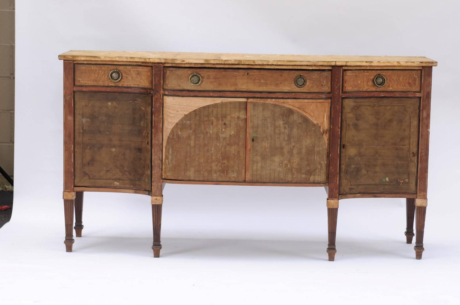 A French neoclassical style stripped wood serpentine four-door sideboard with tapered legs. This French four-door buffet features a serpentine shaped-top over three drawers and four doors. The ensemble is raised on six fluted tapered legs. When we