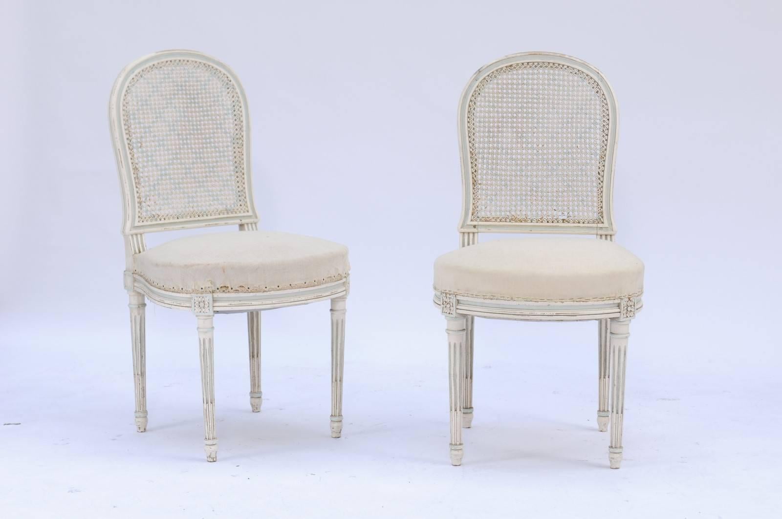 A pair of French Louis XVI style wooden painted dining room chairs with cane backs and upholstered seats from the 1940s. From a trusted vendor in northern France come these lovely Louis XVI-style cane-backed chairs with fluted legs and rosettes on