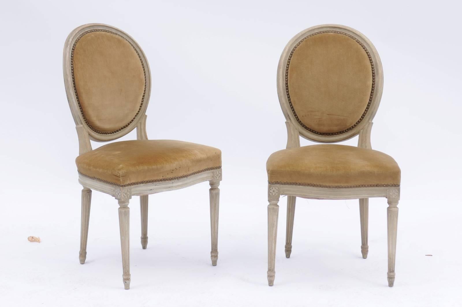 A pair of French Louis XVI style painted wood side chairs from the early 20th century with oval backs, original upholstery, nailhead trim, carved rosettes and fluted legs. We currently have one pair available, priced and sold per pair: $775. The