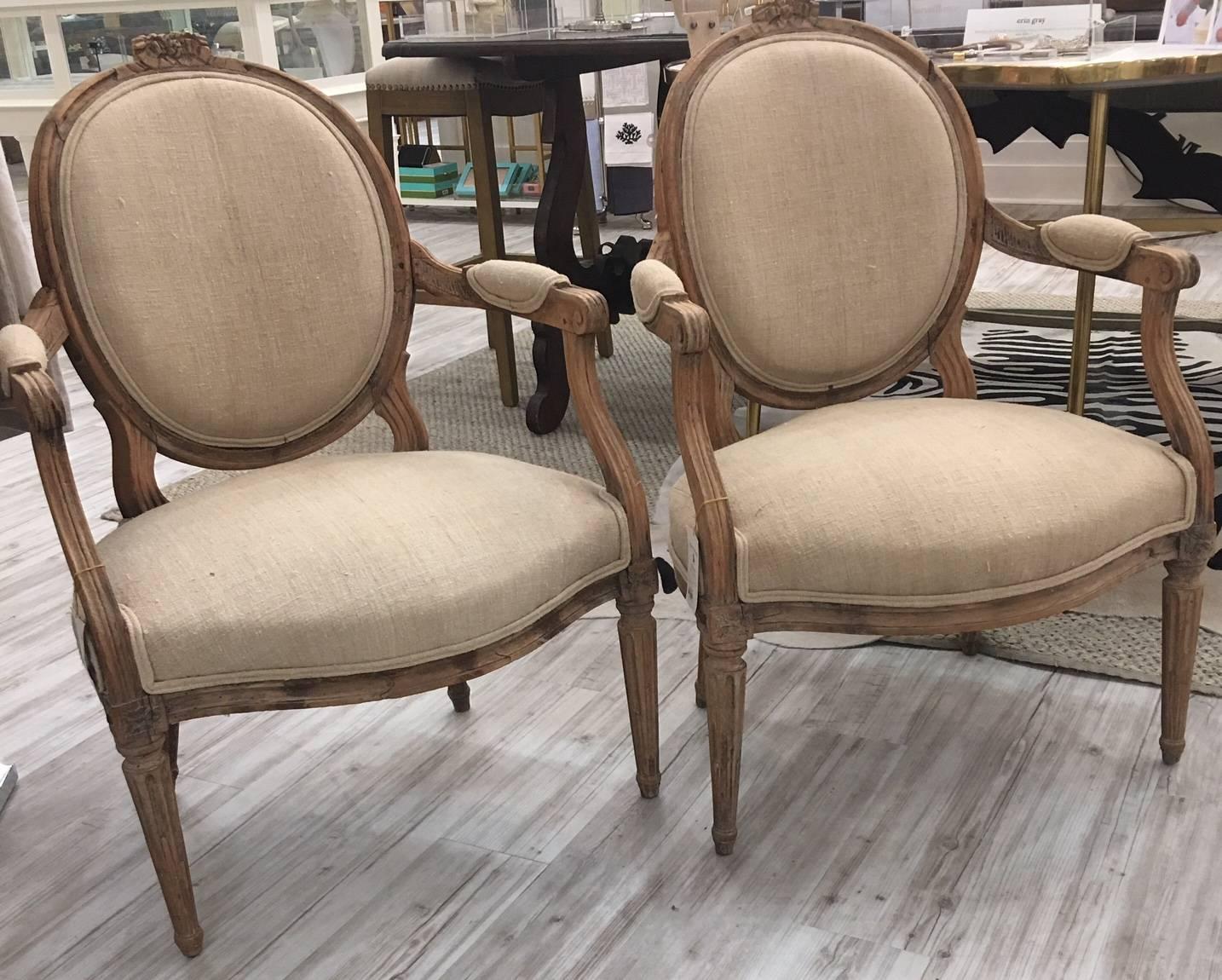 This pair of French, Louis XVI style mid-19th century armchairs is made of carved limed oak. The chairs feature the typical Louis XVI decorative vocabulary. The oval backs are topped with a ribbon carving. The open, partially upholstered arms are