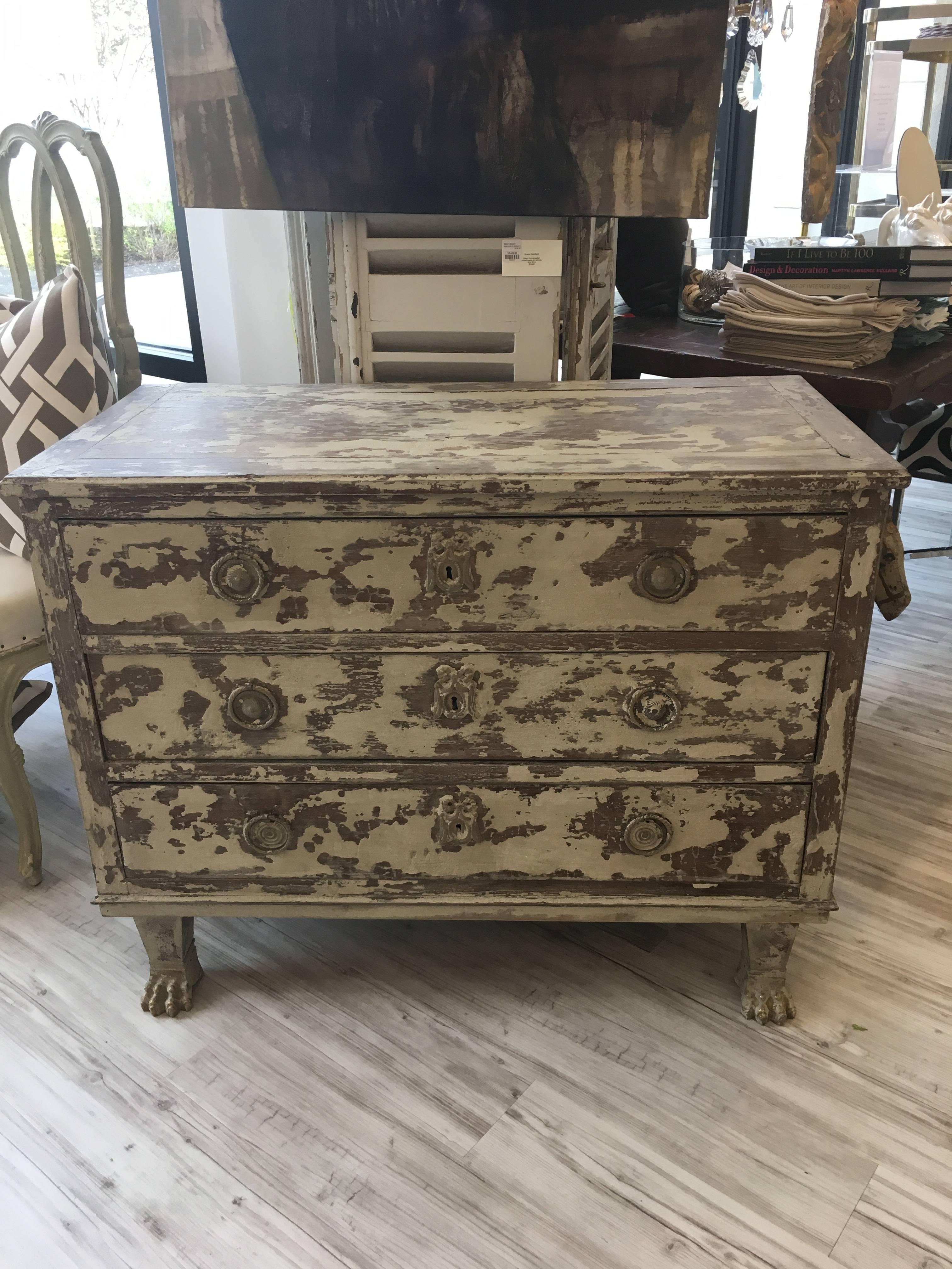 This French distressed finish commode from the early 20th century features a simple rectangular top over three dovetailed drawers. Each drawer is adorned with two ring pulls and a central escutcheon with a Louis XVI knot motif. The sides are made of