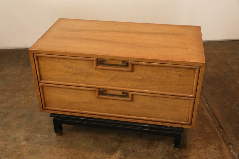 Offered is a Mid-Century Modern American of Martinsville Asian / chinoiserie inspired two-drawer bedside table in walnut with brass pulls and sabots and black wood base. This piece is accented with a sculpted black lacquered base and designed with a