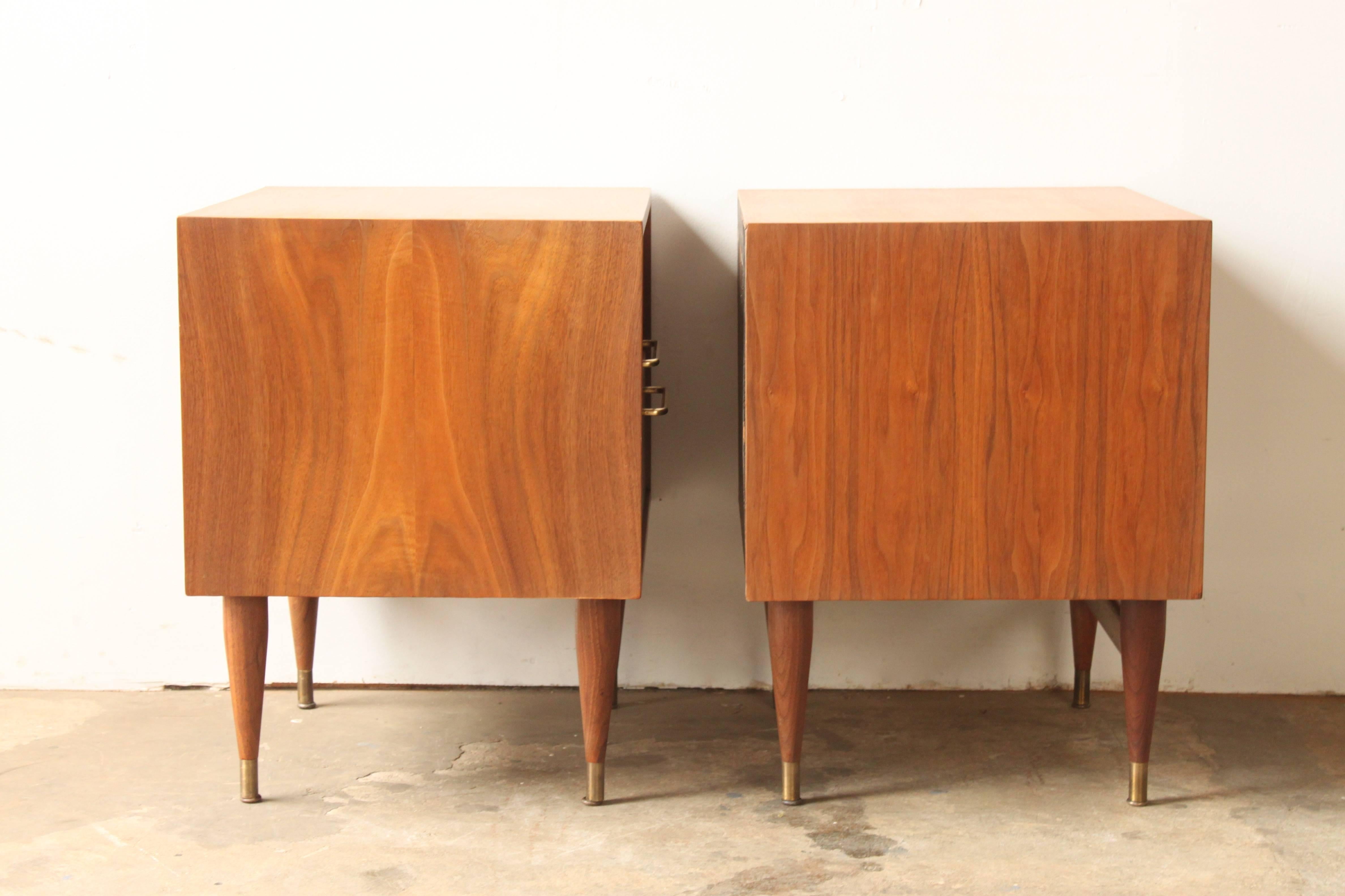 A pair of perfectly executed mid-20th century bedside tables. Rich mellow brown walnut grain is the backdrop for the satin brass drawer pulls and leg tips. Excellent original condition.