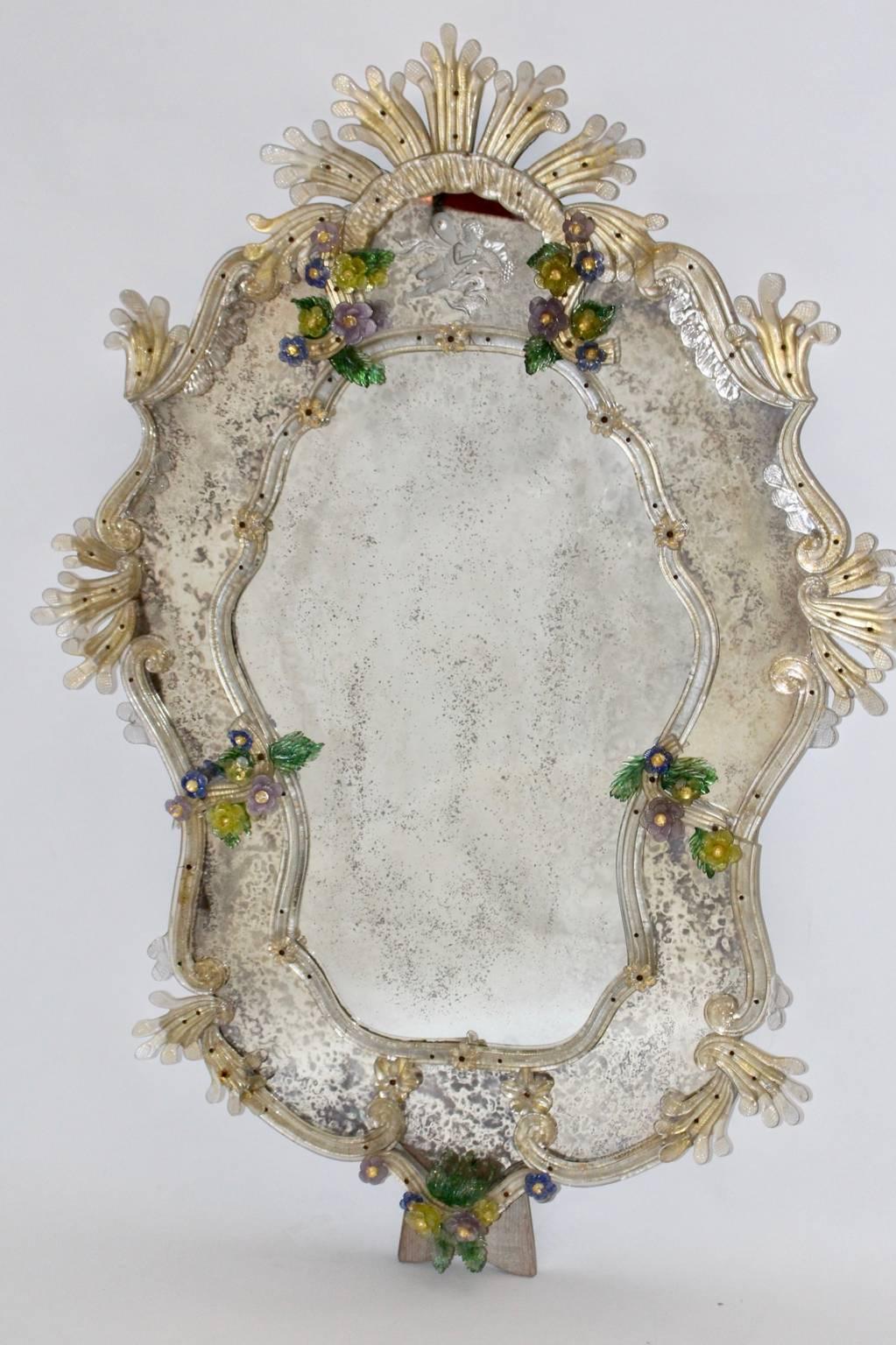 This charming handmade wall mirror was designed and made in Venice in 1950s.
The frame of the mirror is decorated with many multicolored handcrafted Murano glass flowers and elements.
At the upper part of the mirror you see an engraved putto amor
