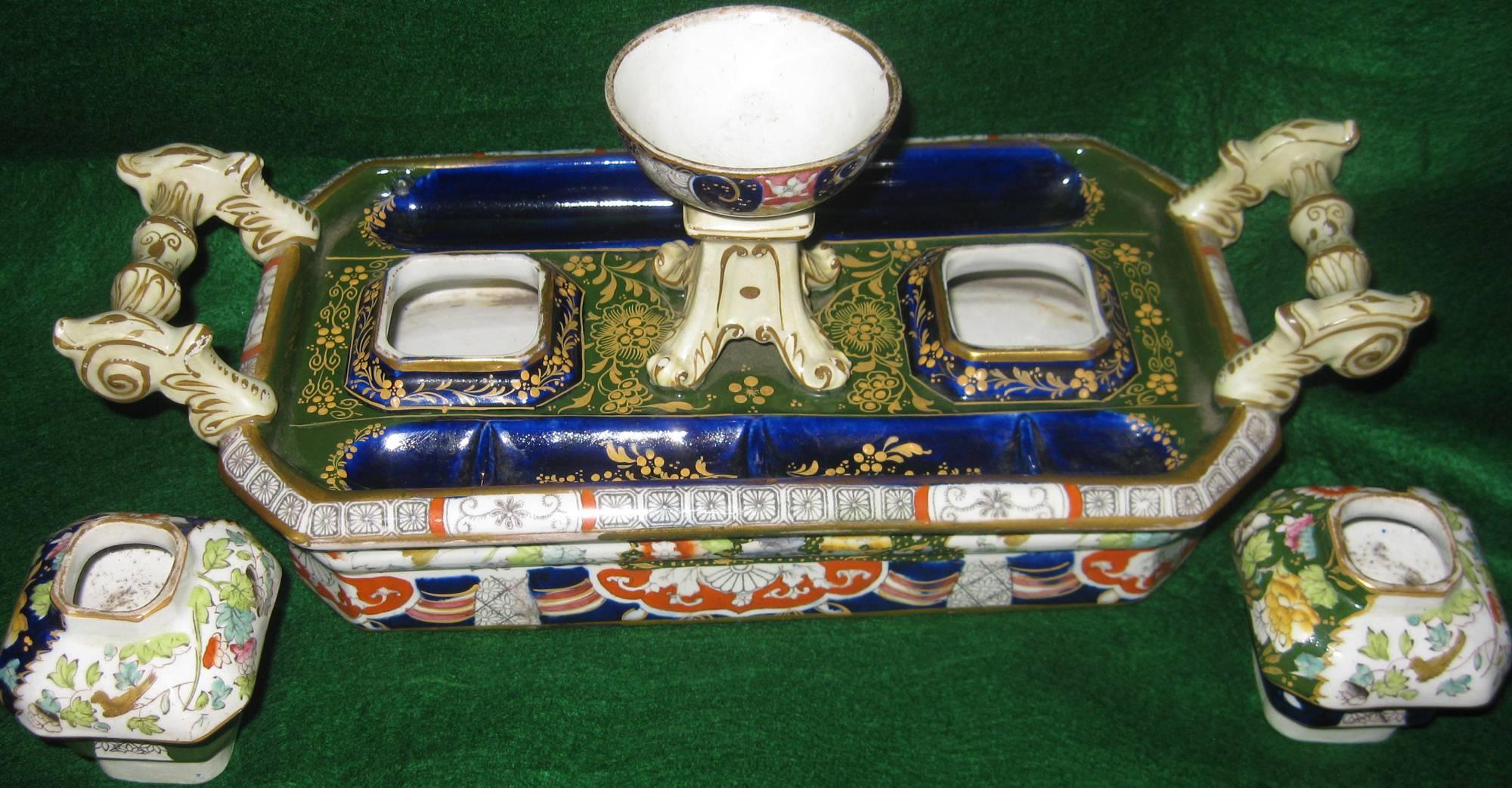 Chamfered rectangular shaped inkstand featuring gilt turned and scroll side handles, two pen trays flanking two raised urn-form inset inkwells and a central circular footed dish. In rich cobalt blue, deep greens, pink and orange with gold accents,