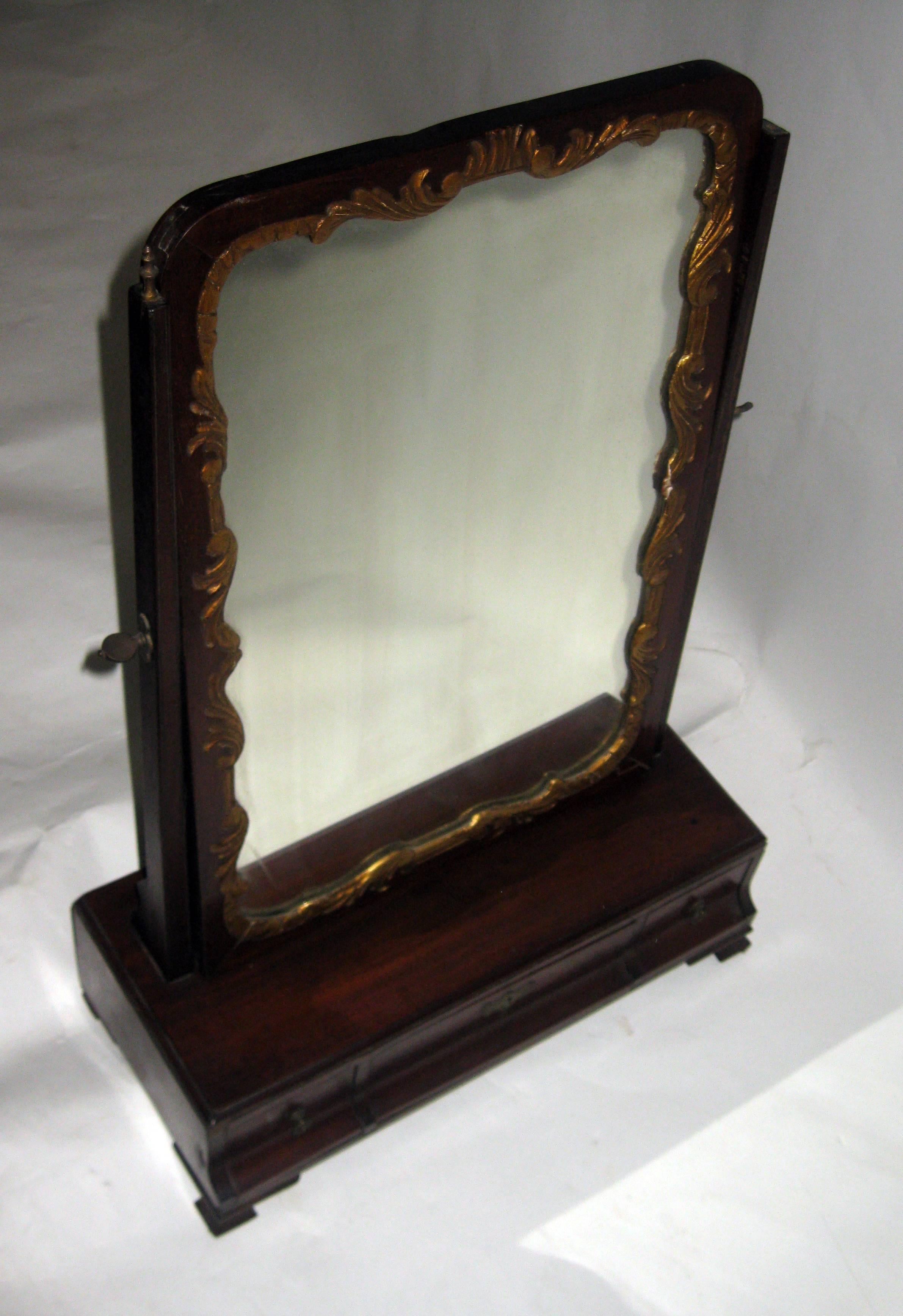 19th century Georgian mahogany dressing or shaving mirror featuring original brass hardware, three drawers and ogee bracket feet. A.fancifully carved giltwood frame surrounds the swivel mirror that has a wooden back. See measurements below.