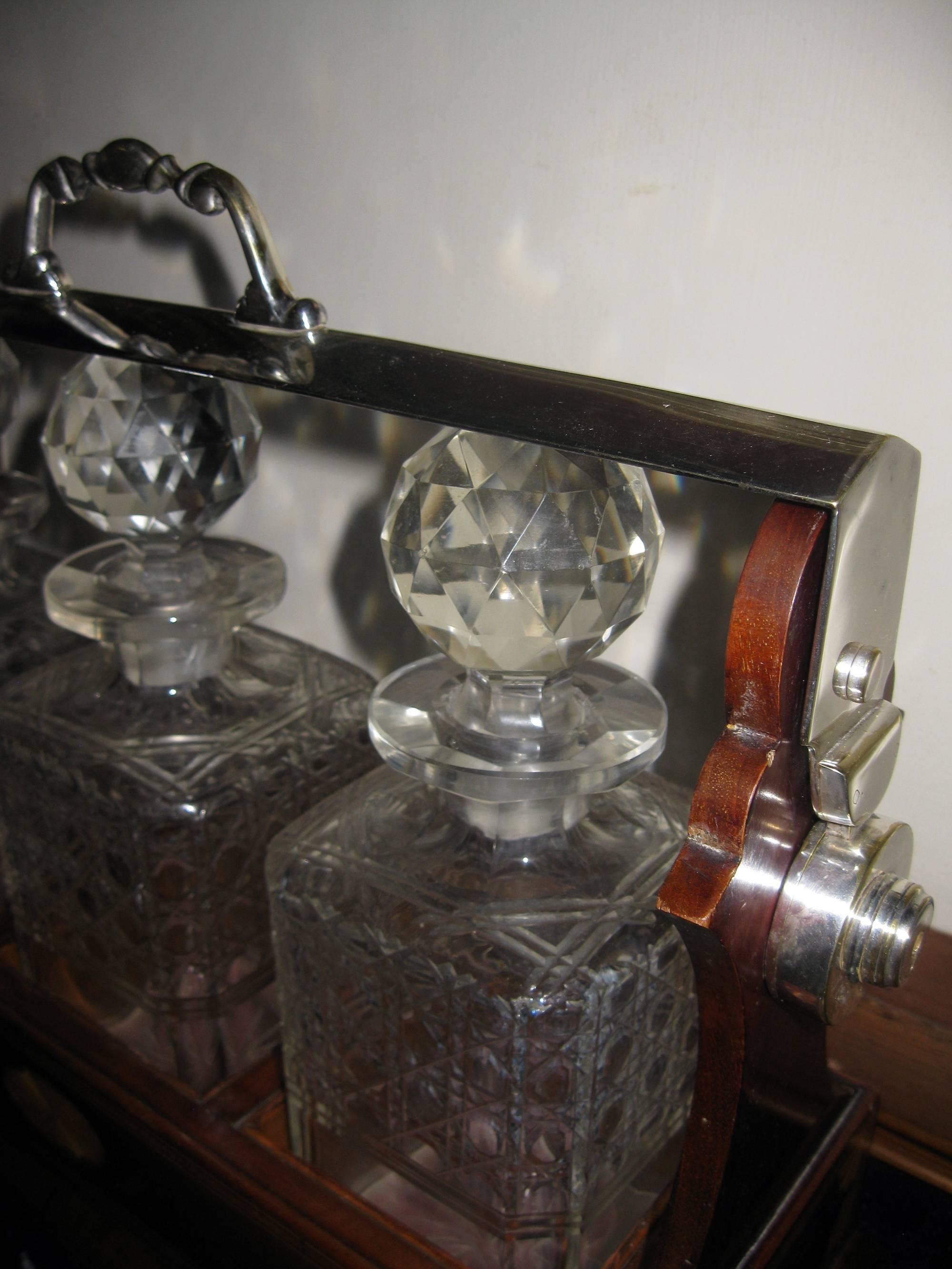 English mahogany three decanter Tantalus featuring silver handle and locking mechanism top. The original cut-glass decanters with stoppers are in a fitted case with cross band inlay on panels with corner and centre shell medallions. Exquisite