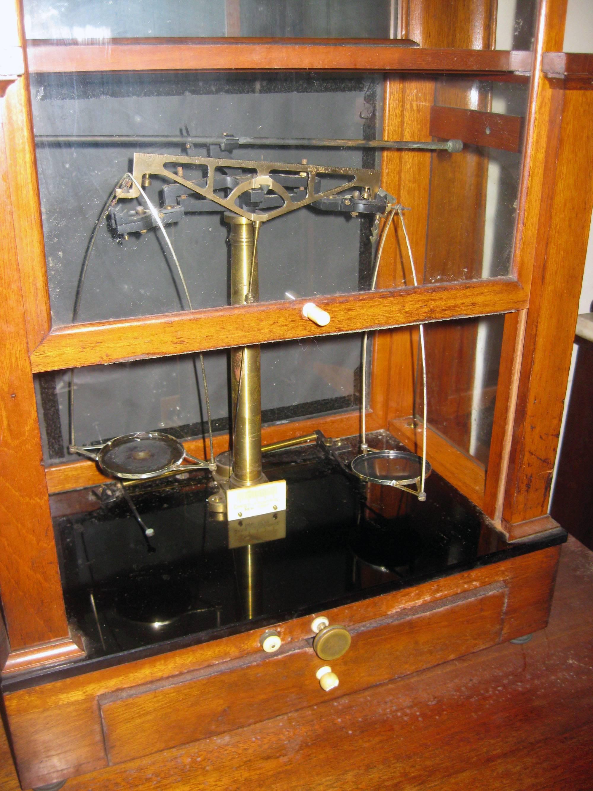 Double balance scale featuring a mahogany wood case, black glass platform, lift front glass side, and a bottom pull out drawer. Fitted with a level. Brass screw-on feet. Bone and brass pulls. Label reads Christian Becker Inc. New York. They were
