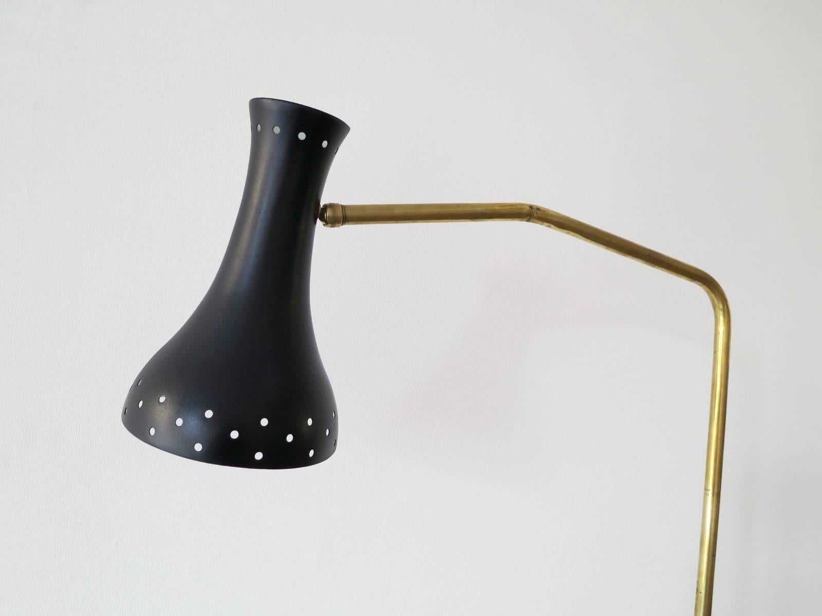 A versatile floor lamp in brass with a black painted and perforated shade. The base in cast iron with a segmented wooden base cover. The brass stem on a pivotable ball joint allowing a wide range of movement and also telescopic stem for height