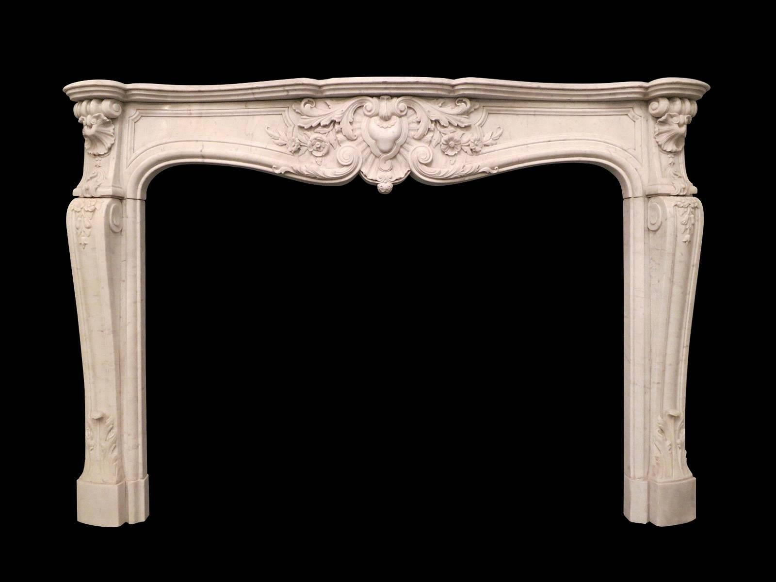 A Louis XV style fireplace in pale Carrara marble, the frieze with a carved central cartouche of C scrolled foliage, flowers and medallion. The end blocks with carved shell and foliate carvings descending into conforming carvings on the jambs with