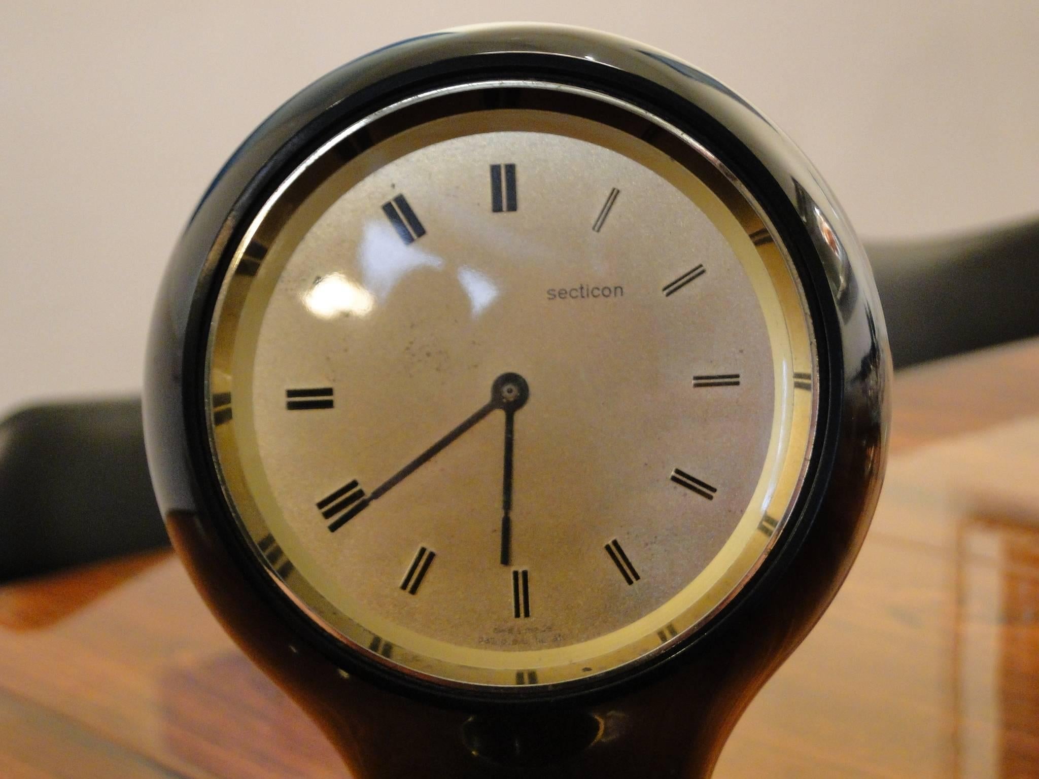 Space Age shape in original black finish. Brass accent details and original Lucite cover.

Stamped with makers label, made in Switzerland.
These desk clocks were manufactured in la Chaux de Fonds, Switzerland.
Measure: Dial Ø 9 cm.
 