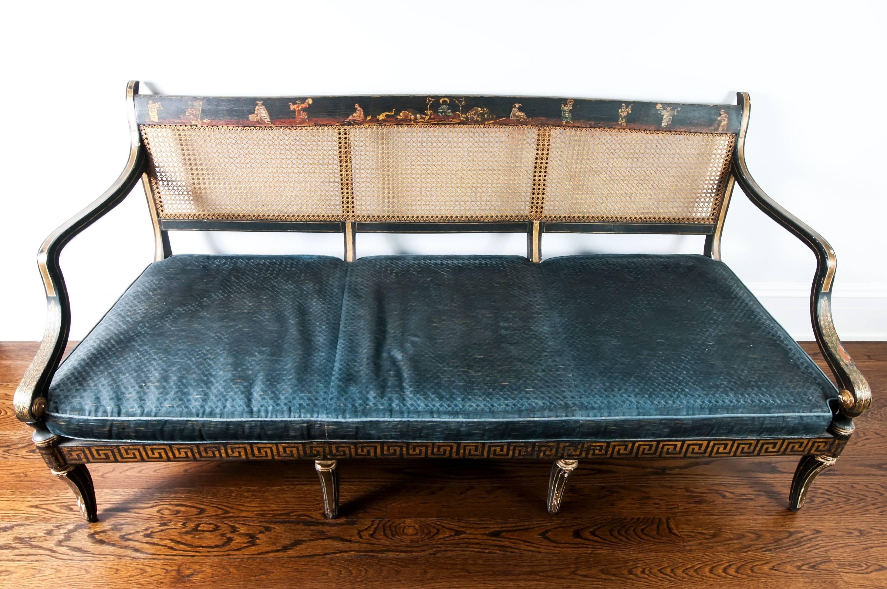 An antique painted Regency caned settee with velvet cushion. Frame is painted black with gilt accents and with chinoiserie imagery, the back and seat are caned, with the caning fully intact. The velvet cushion is original and shows overall wear. 