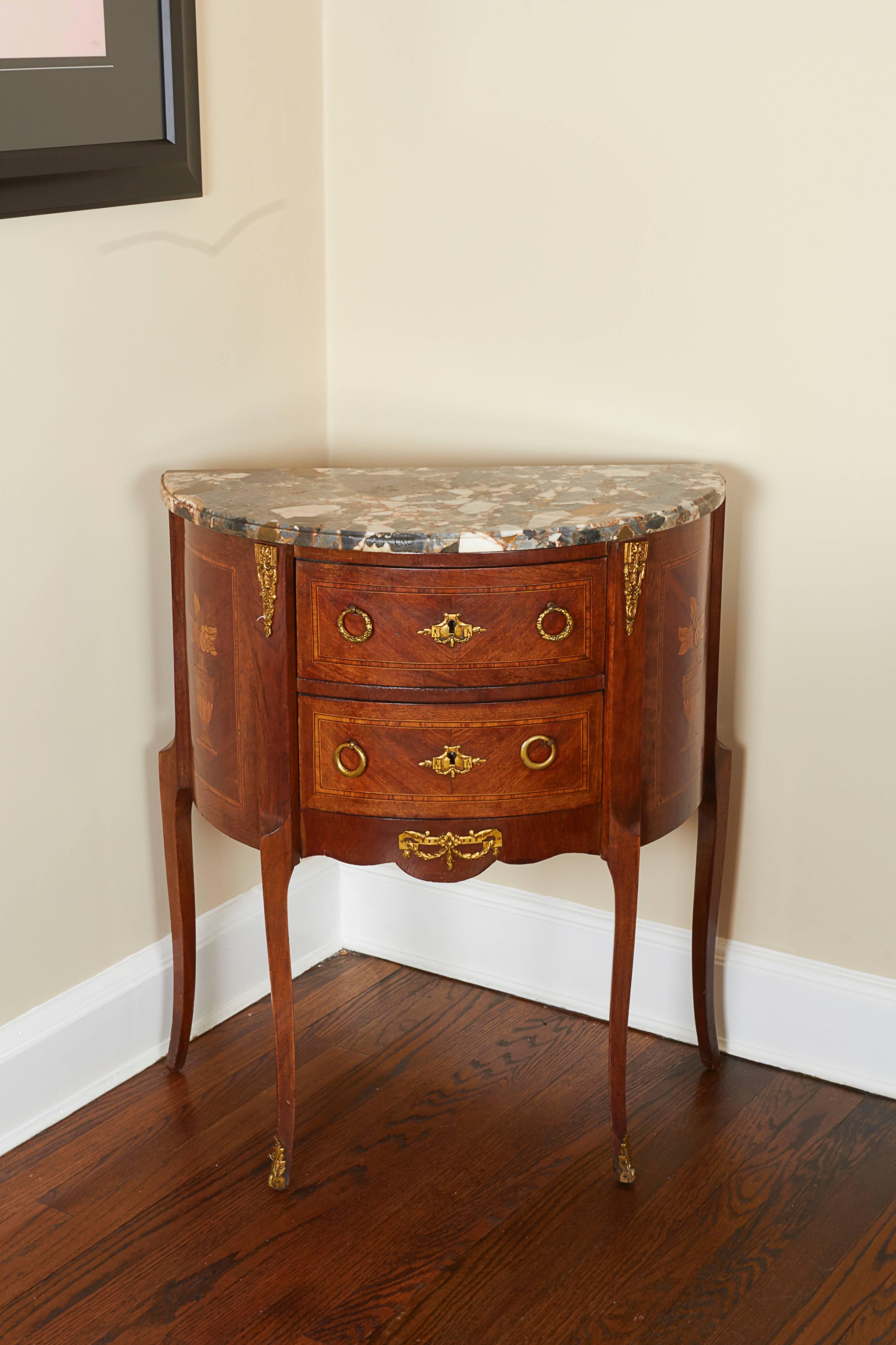 Each small scaled commode feature a demilune marble top over two bookmatched veneers drawers mounted with laurel wreath ormolu pulls, and inlaid with flower-filled Classical urns to the sides; raised on cabriole legs terminating in sabots.