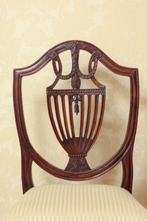 A fanciful Italianate interpretation of the Classic shieldback chair. Featuring foliate carving with a pierced urn shaped splat. The drop-in upholstered seat over a chevron-carved rail raised on square tapering, reeded legs joined by an H-form