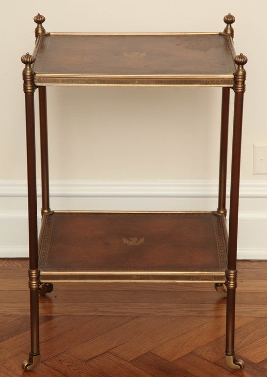 The table inset with gilt-tooled in brown leather. The rectangular brass bound top and undertier on tubular legs with finials supported by brass feet terminating in castors.