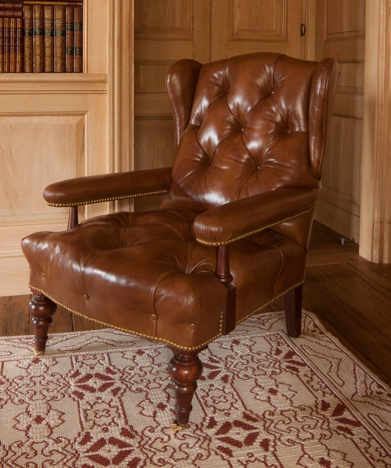 Handsome pair of generous but still elegant scaled armchairs covered in a deep toffee-covered leather with brass nailhead trim; featuring subtle wings to the back and mahogany turned legs on brass caps and casters.