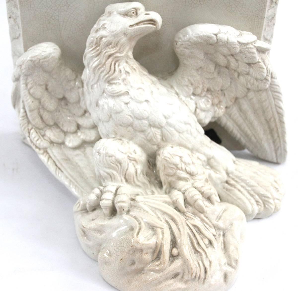 Pair of early 19th century Federal period American eagle form wall bracket shelves in white glaze, retaining the original metal tab hangers, in non-opposing stances, standing atop a rocky outcrop with sheaf of grass grasped in talons, supporting a