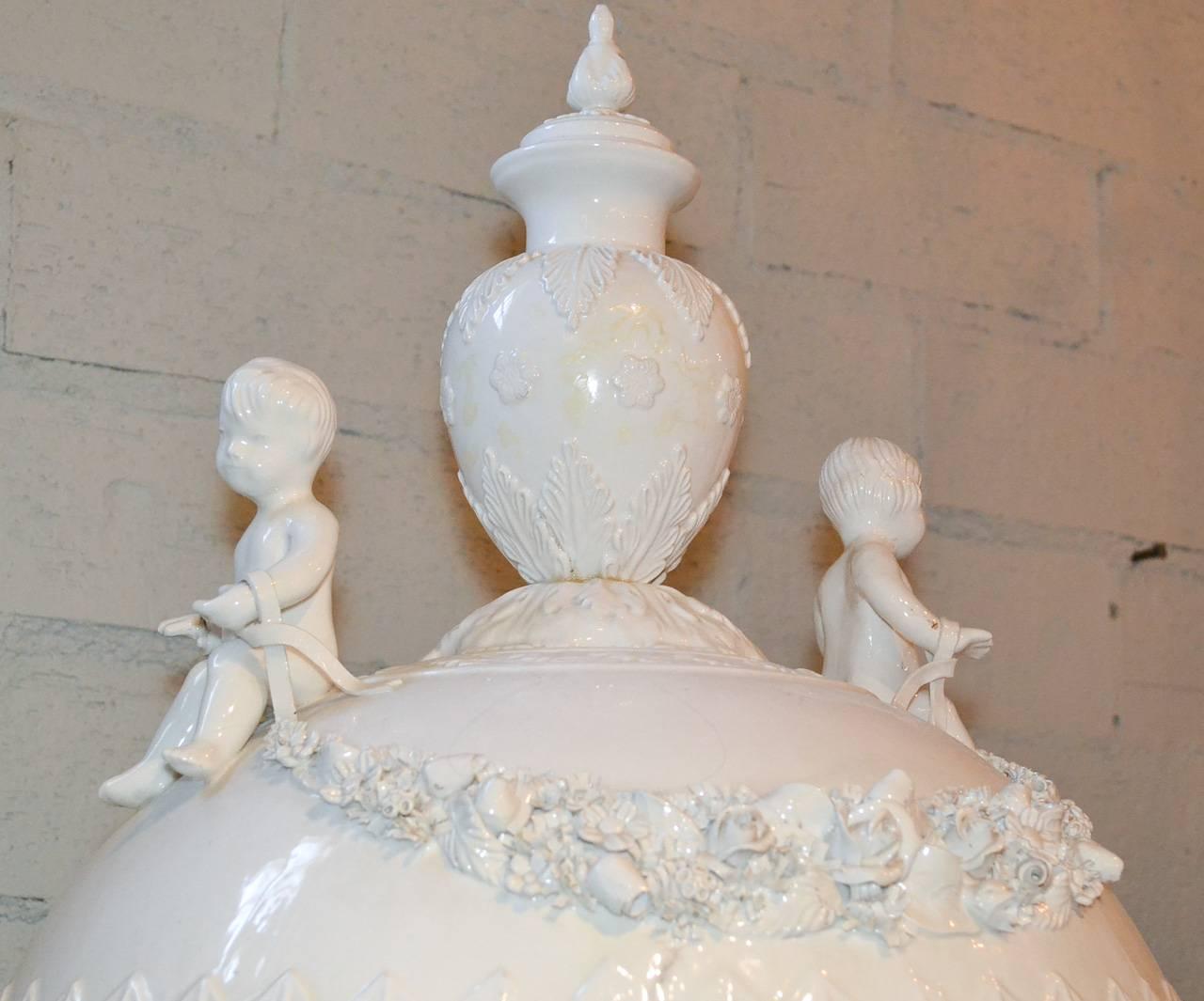 Stunning pair of large Italian glazed porcelain capped urns. Having beautifully intricate floral swag detailing, charming putti adornments, and wonderful glazed patina. Beyond chic for numerous designs!