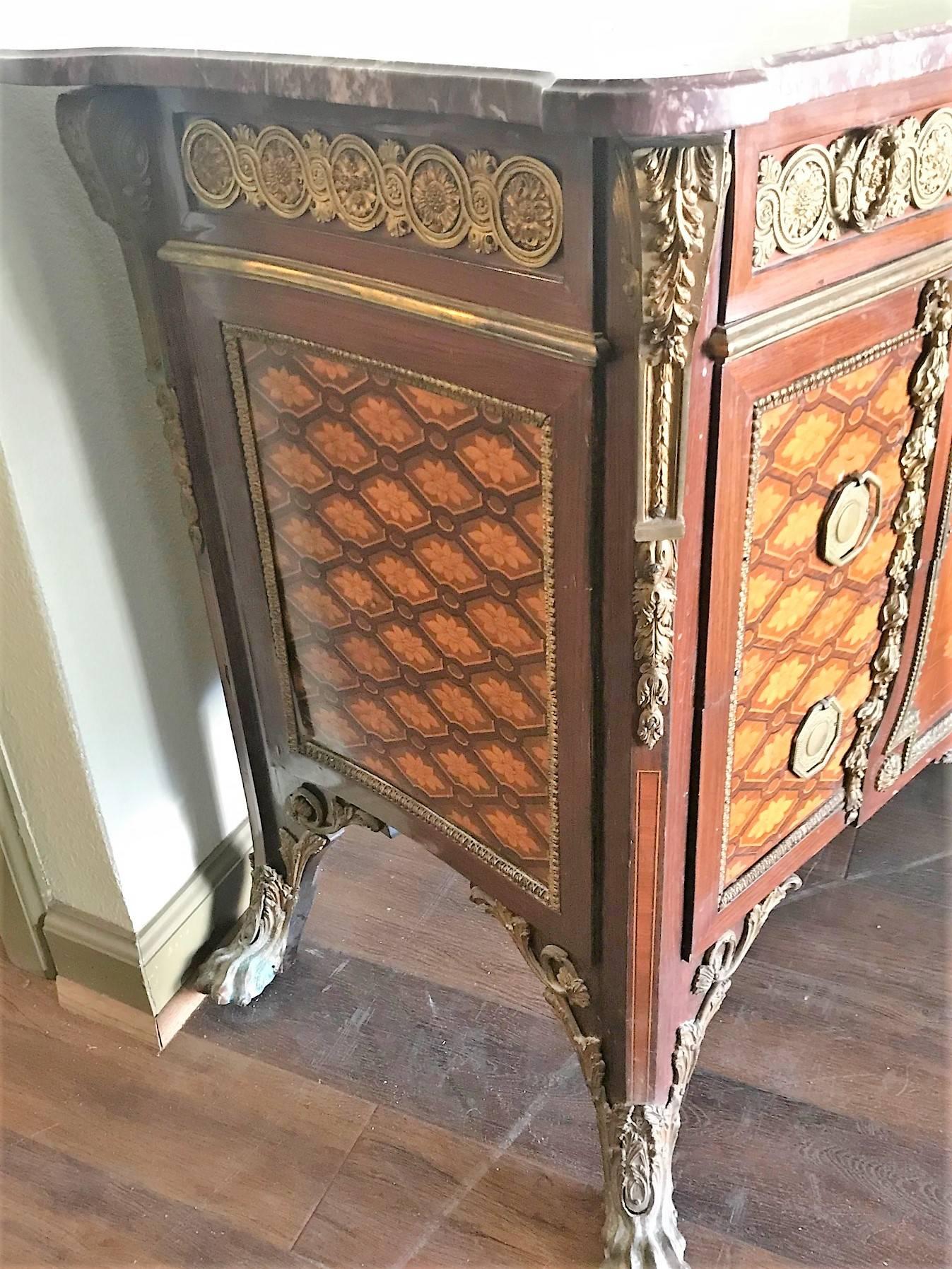 Superb circa 1930s French transitional style commode with a shaped marble-top.

The frieze ornately decorated with gold-gilded bronze leaf scrolls above a central panel with marquetry inlays of exotic woods depicting a large urn with flowers and