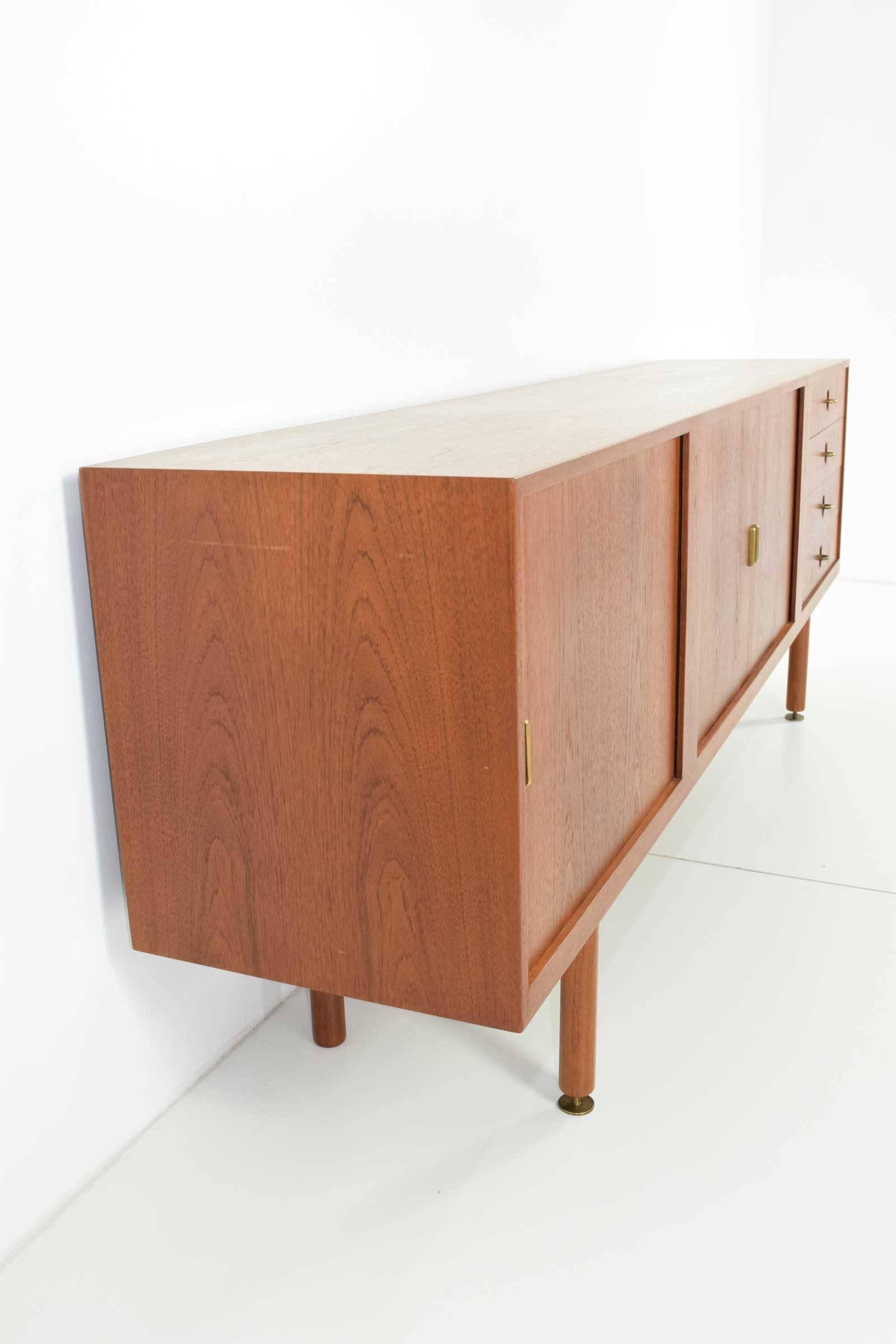 This is a gorgeous credenza/sideboard in teak. Has three tambour doors that recess into credenza when open, interior is fully finished as well as back so the piece can float in a room if desired. Hardware is solid brass, decorative leveling feet on