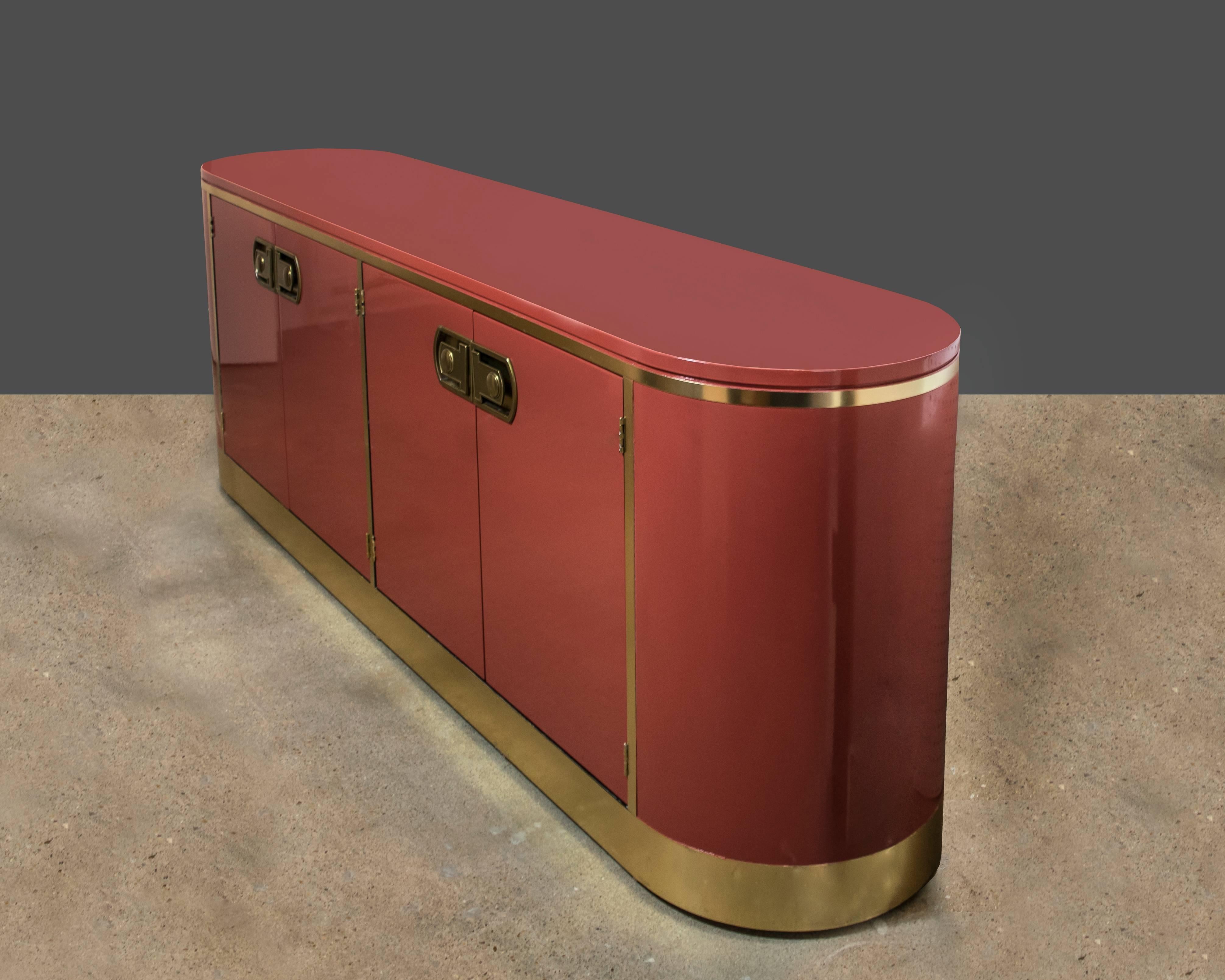 A breathtaking brass and Moroccan red lacquer credenza manufactured by Mastercraft Furniture Co. It is extremely well designed and has rounded corners with four cabinet doors and storage shelves inside.