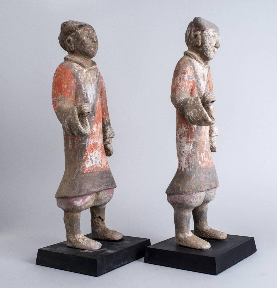 Chinese Han dynasty pottery funerary figures, each with remnants of orange glaze decoration, both with one hand raised mounted on removable bases.

Dimensions: 18