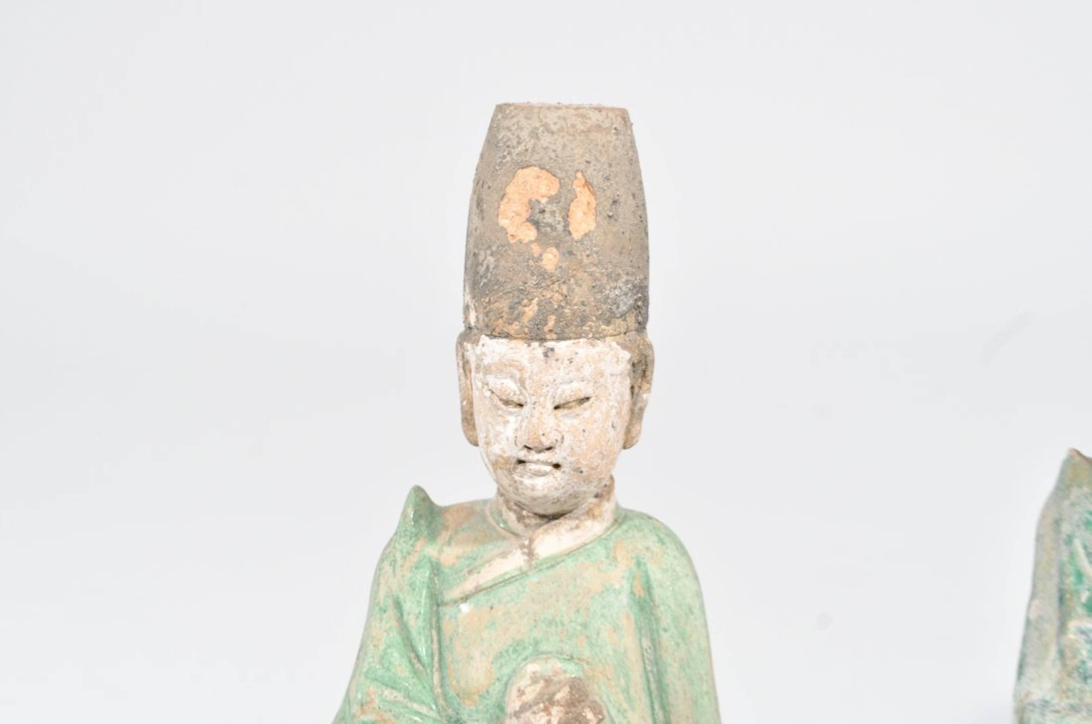 A pair of Chinese male earthenware attendant figures, hand-painted with pigments. Both figurines wear a tall black cap, green robes. Both figures are hollow and Stand on a raised hexagonal base. Figures such as these often were buried with the