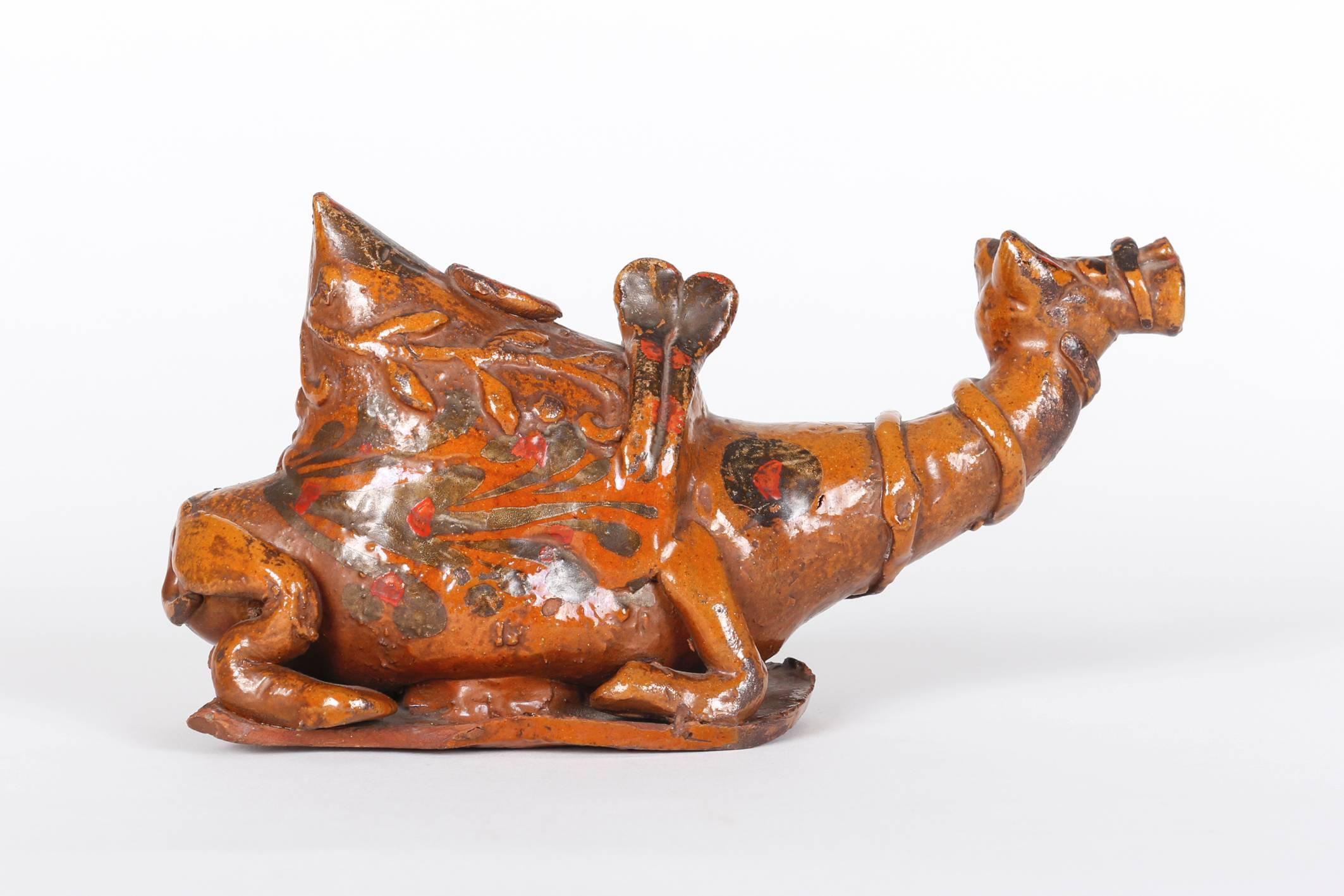 A kneeling camel with a saddle, its neck stretching out front, set on an oval plate. Red earthenware with a yellow glaze. Strikingly decorated on the back with a leafy branch in relief, the same motif being repeated on the bottom in white paint over