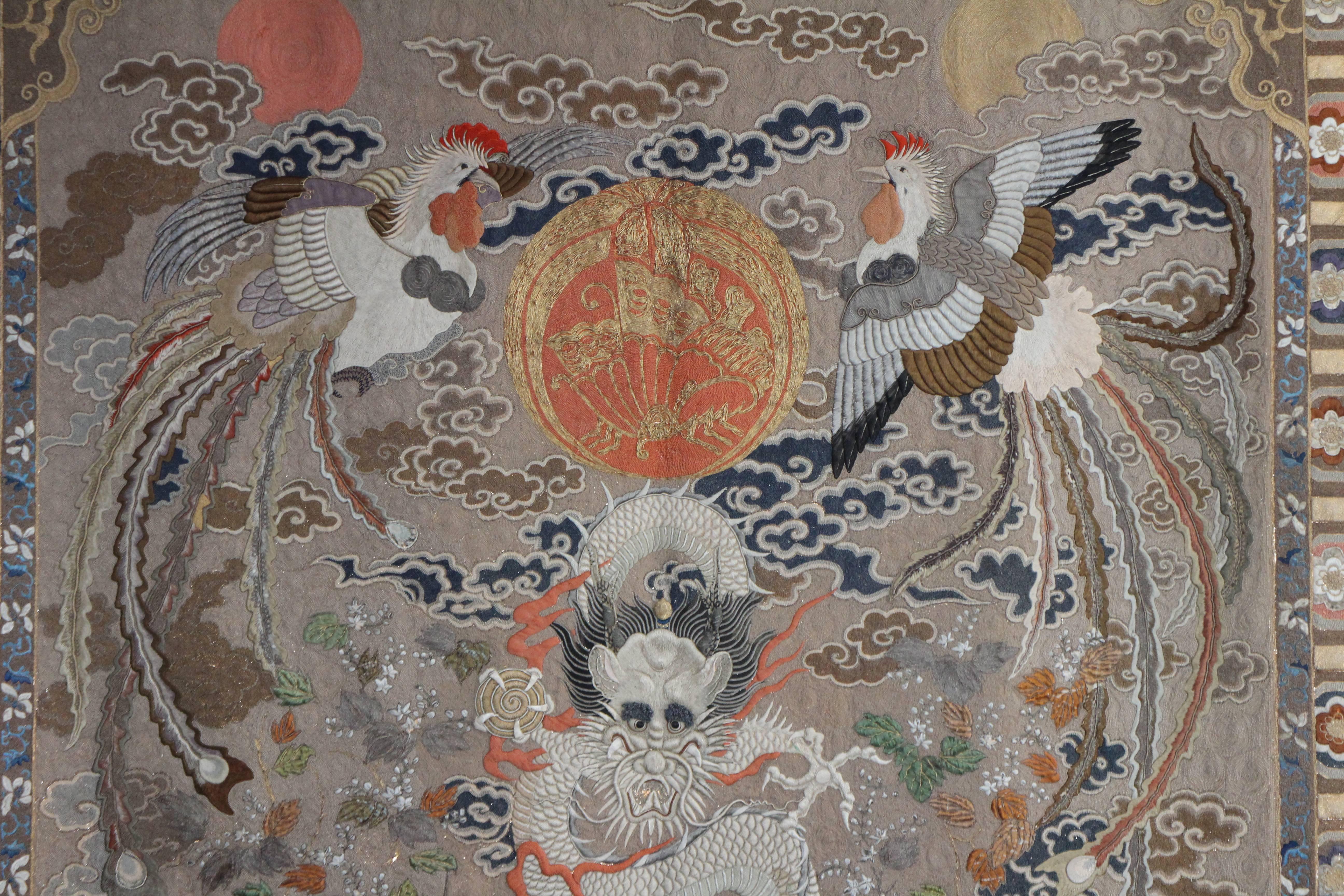 An impressive antique silk embroidery of a five toed dragon (traditionally associated with imperial China) flying over a turbulent sea and rolling hills with galloping stylised horses. Above the dragon two phoenixes flank a variation of the Taira