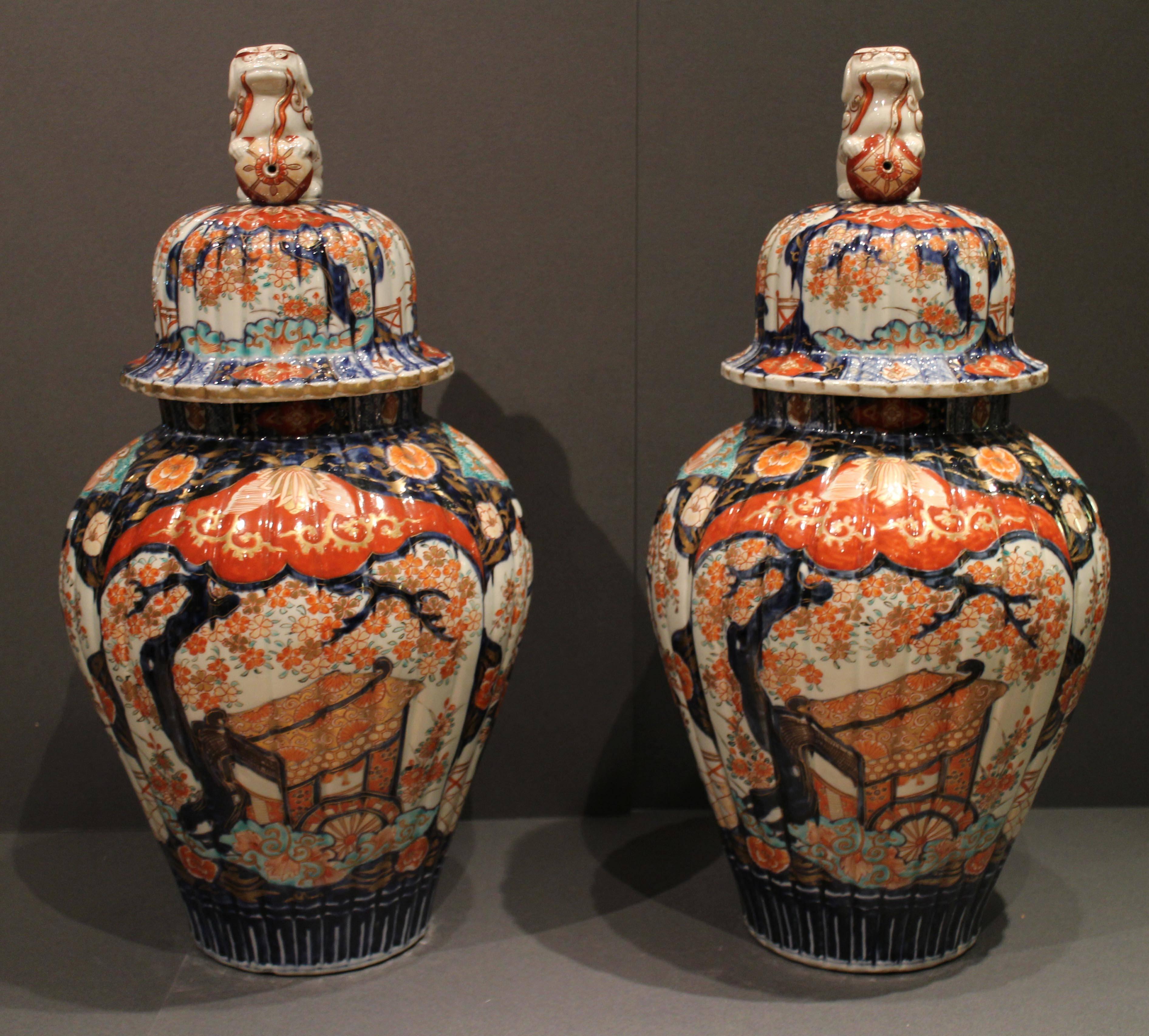 A pair of antique Japanese Imari vases and covers decorated with large shi shi dogs with balls, the vases are decorated in blue and orange with traditional Japanese scenes of flower arrangements and flowering blossom trees.