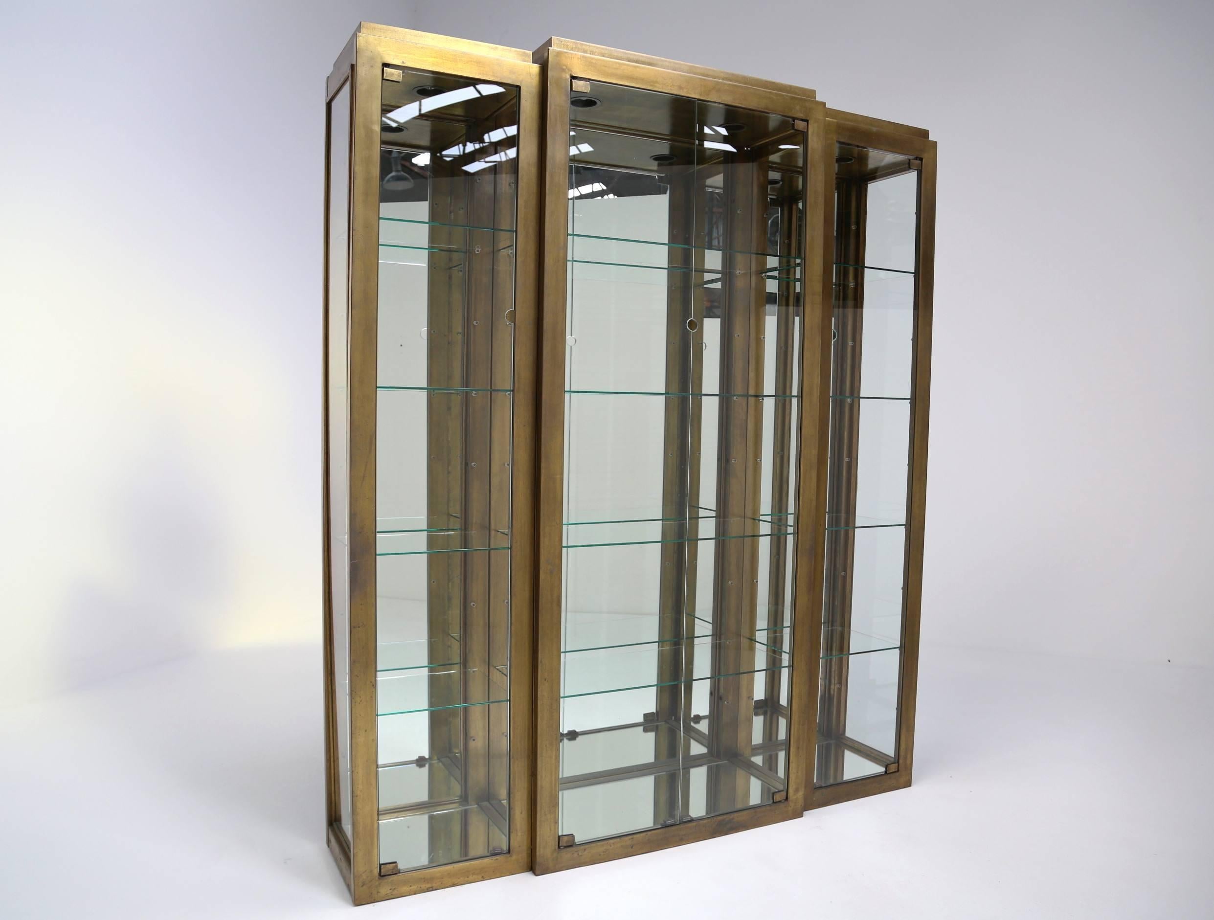 A plated brass Art Deco style four-door display cabinet with internal lighting. Two narrow side cabinets attach to a larger central cabinet to form a large stepped Art Deco style vitrine. In beautiful original condition and a spectacular piece for