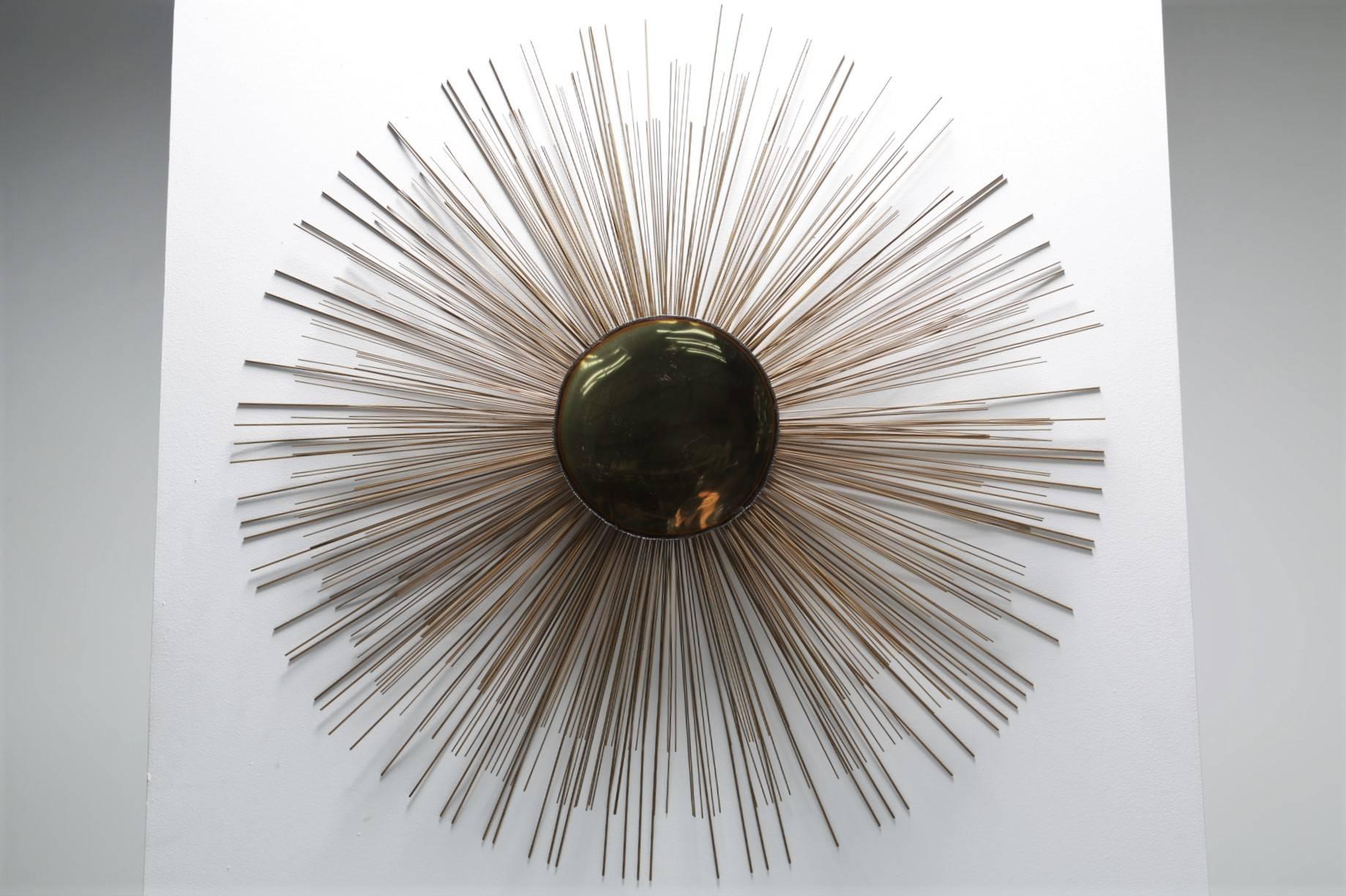 This is an impressive sunburst wall sculpture by design partners Curtis Jere or C. Jere. Large in size, this piece evolves around a shiny brass centrepiece. This is then edged in metal and has metal rods emanating from it, much like a sunburst.