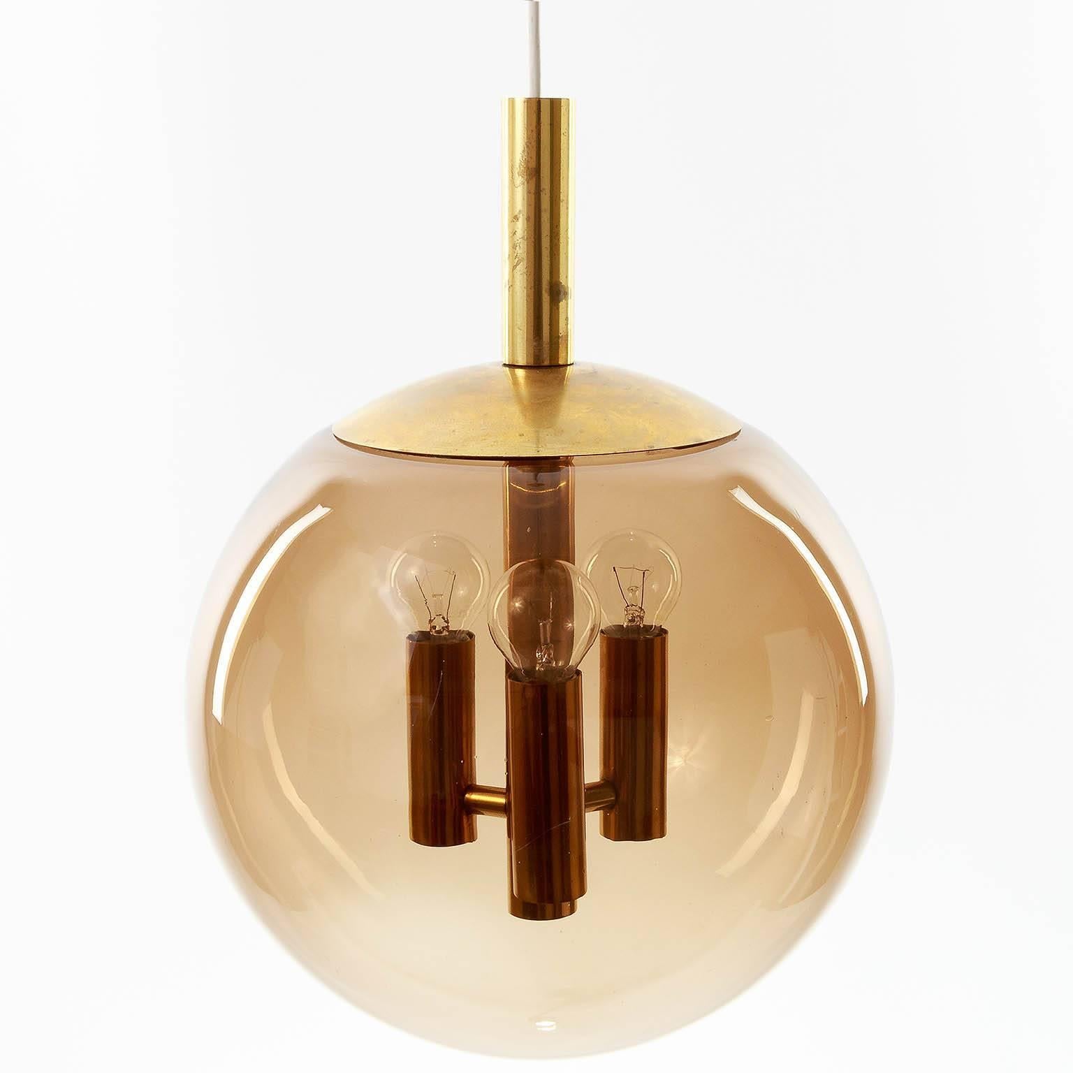 An amber tone or smoked handblown glass globe pendant light by Limburg, Germany, manufactured in Mid-Century, circa 1970 (1960s or early 1970s). A polished brass fixture with little and nice patina. 
The lamp takes three small base bulbs. It