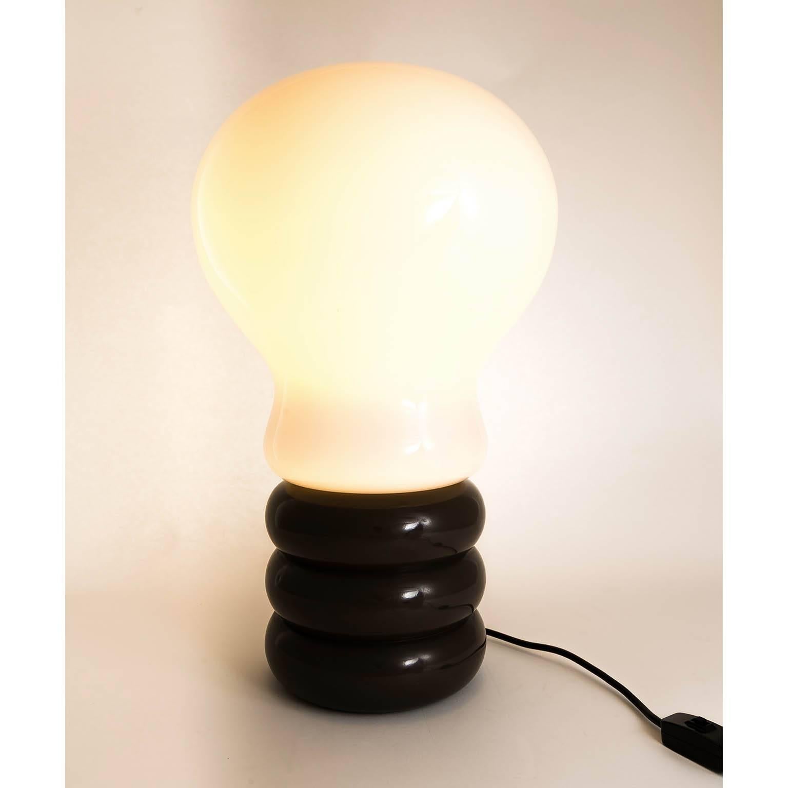 The bulb table lamp was designed by Ingo Maurer and produced by M-design in Munich in the 1970s. 
It is made of a dark brown metal stand and a blown opal glass lamp shade in the form of a bulb.
There is one socket for a medium or standard screw base