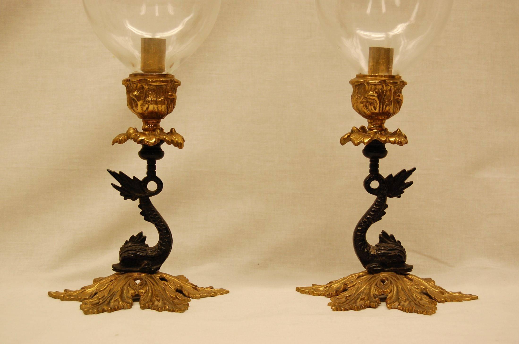 Pair of candlesticks with large heavy glass hurricanes on brass bases with dolphin form stems in black paint.