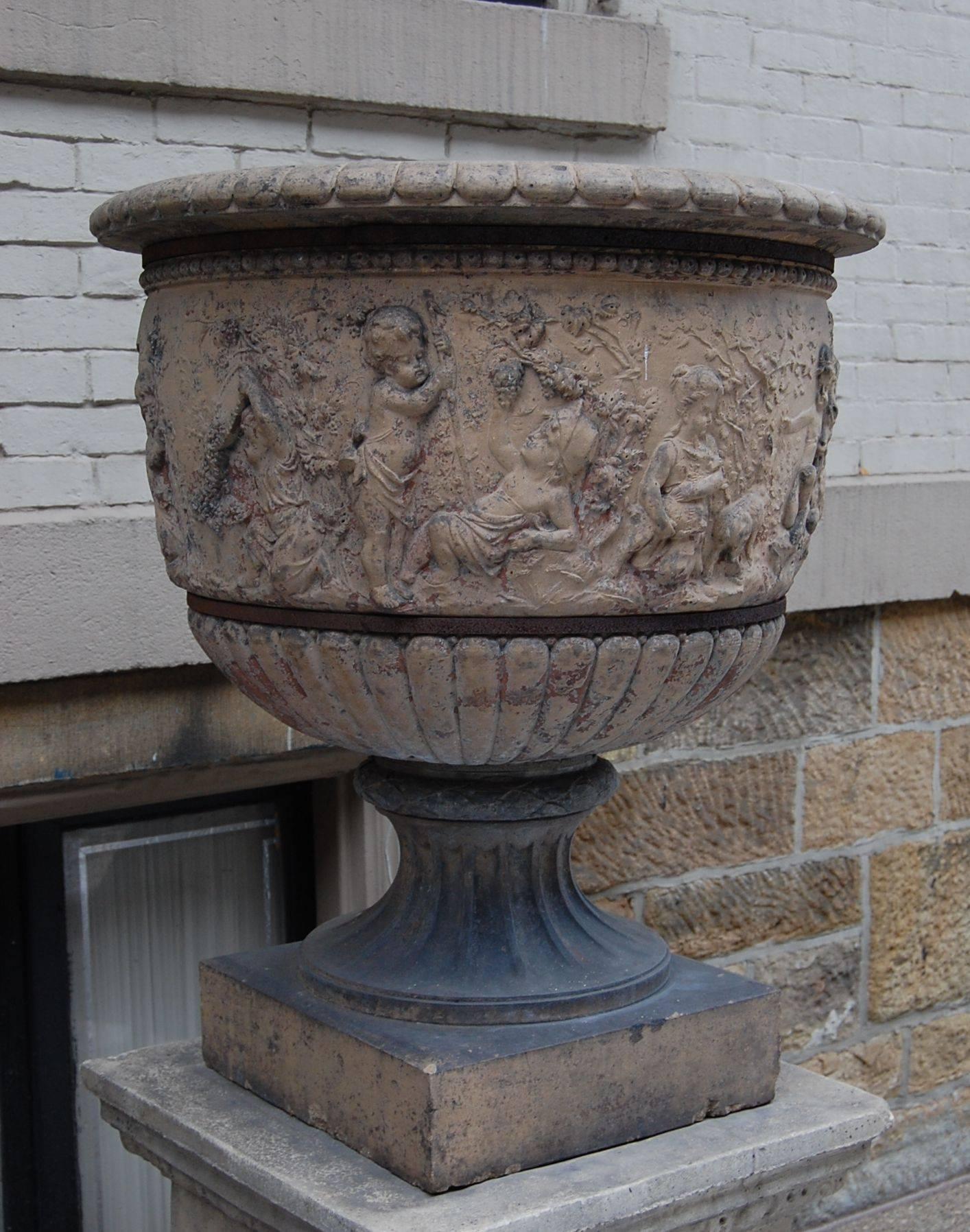 This large terracotta urn shows playful children and goats frolicking, as well as continual grape vines and trees around the entire perimeter. It has two steel retaining rings mounted to secure the urn and to keep the existing stress crack from