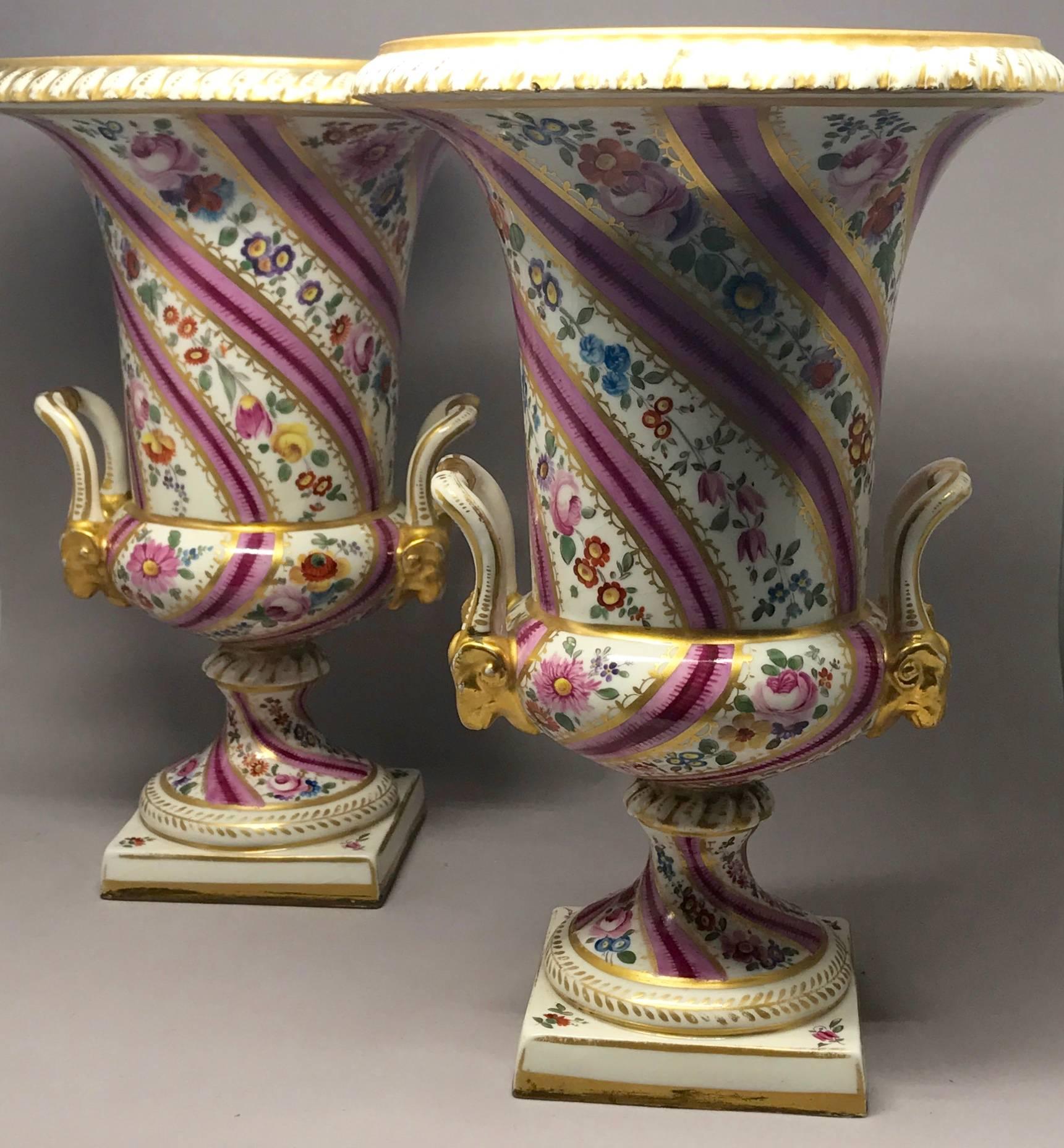 Pair pink floral and gilt porcelain painted urns. Pair urns with vibrant pink, mulberry and fuschia banding alternating with rich floral sprays on the two part body with rams' head handles. In the manner of Swansea, possibly made by Edmé Samson.