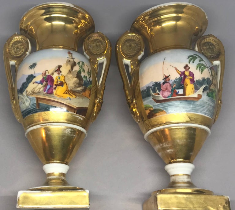 Pair of Empire gilt chinoiserie vases. Pair hand-painted and gilt French vases of the Empire period with mask handles and gilt surrounds centering on richly colored oriental scenes with figures and pagodas in landscapes in yellows, greens and