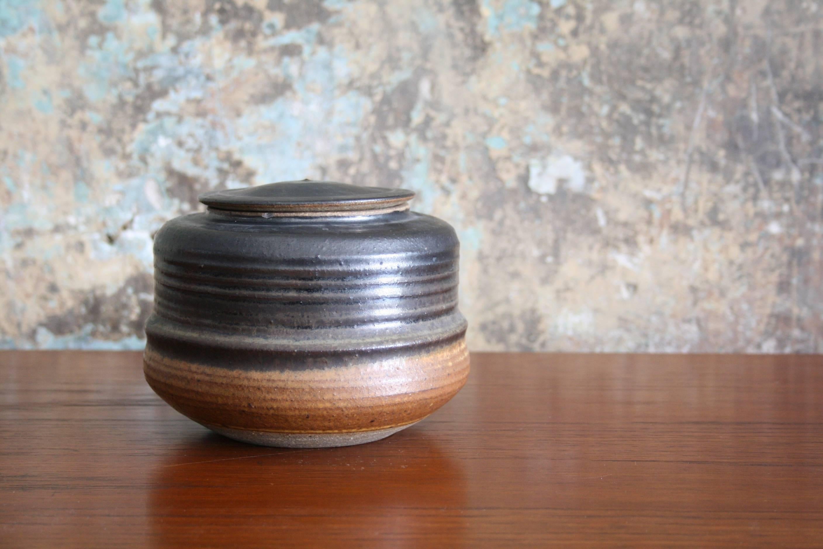 
Karen Karnes (American, 1925-2016) Stoneware ceramic lidded vessel. 
Marked with artist's stamp on side of piece.

 This round vessel has a lightly ribbed texture and a slightly convex, removable, fitted lid. The colors of the smooth glaze
