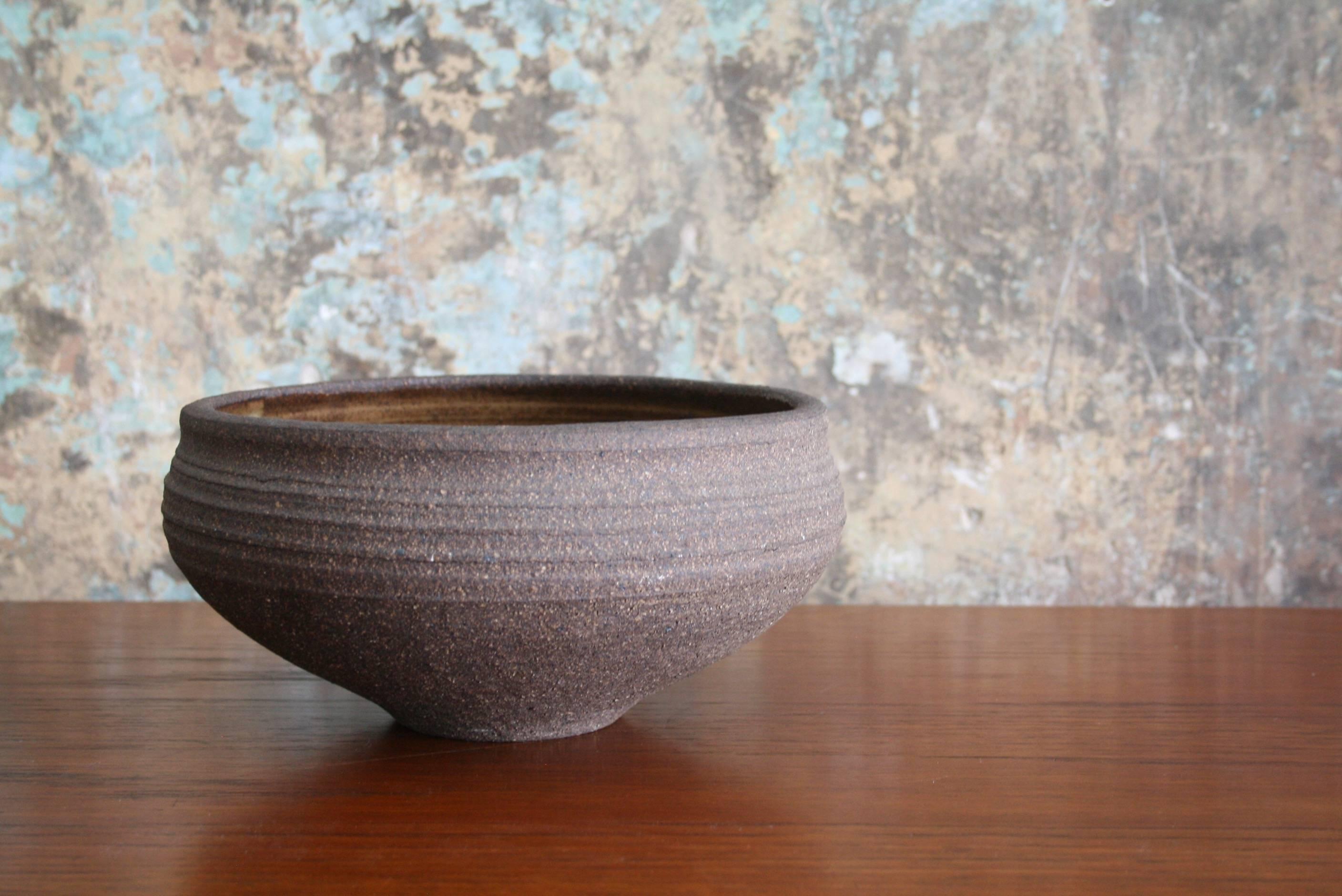 A functional hand thrown stoneware bowl by highly regarded artist Karen Karnes (American, 1925-2016). The wheel-thrown bowl features a glossy mottled eggshell liner glaze, a rough exterior surface with a rich, deep brown earth color, and a mild