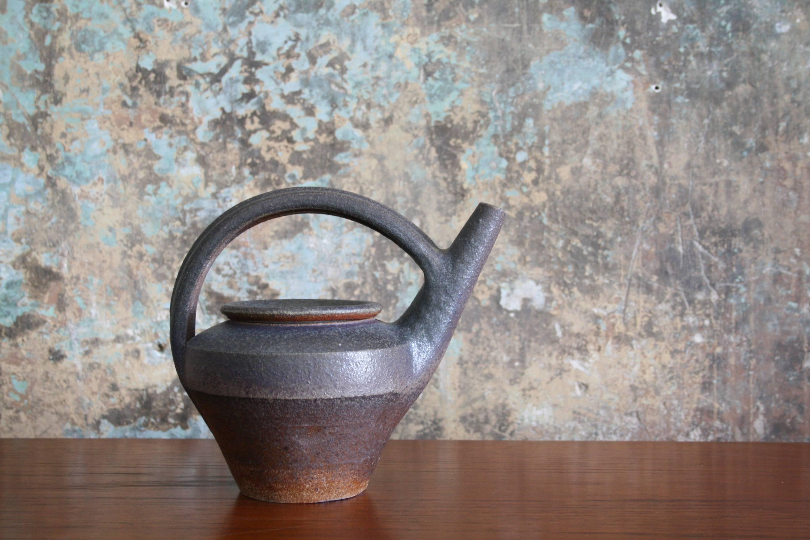 A hand thrown, lidded, functional stoneware teapot by highly regarded ceramic artist Karen Karnes (American, 1925-2016). This architectural orange and brown teapot features an overall rough texture, triangular body shape, and a semi-circular handle