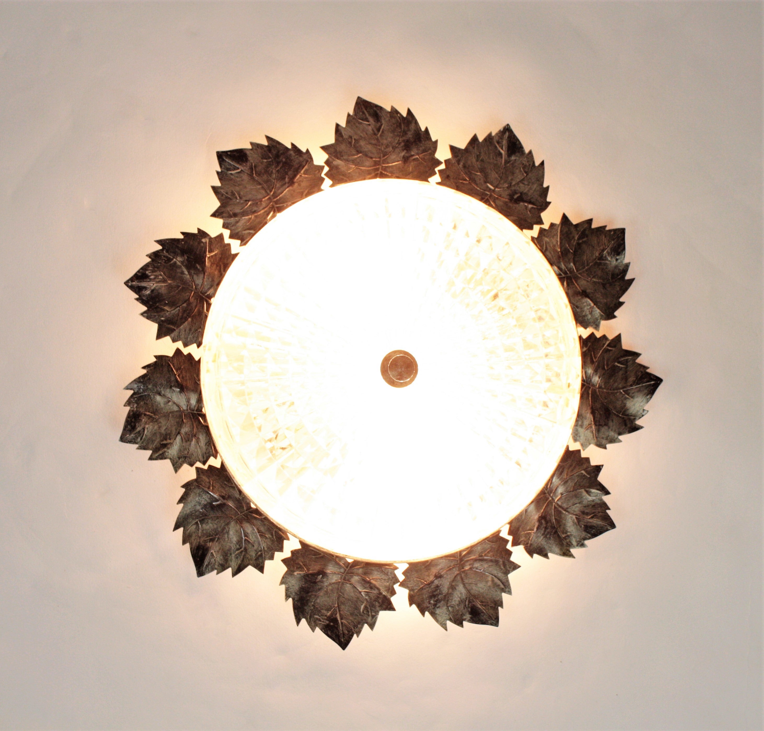 Beautiful ceiling flush mount with iron leaves frame and pressed glass shade. Manufactured by Ferro Art. Spain, 1960s.
This midcentury ceiling light has an iron structure with a ring surrounded by detailed iron leaves patinated in silver-grey color.