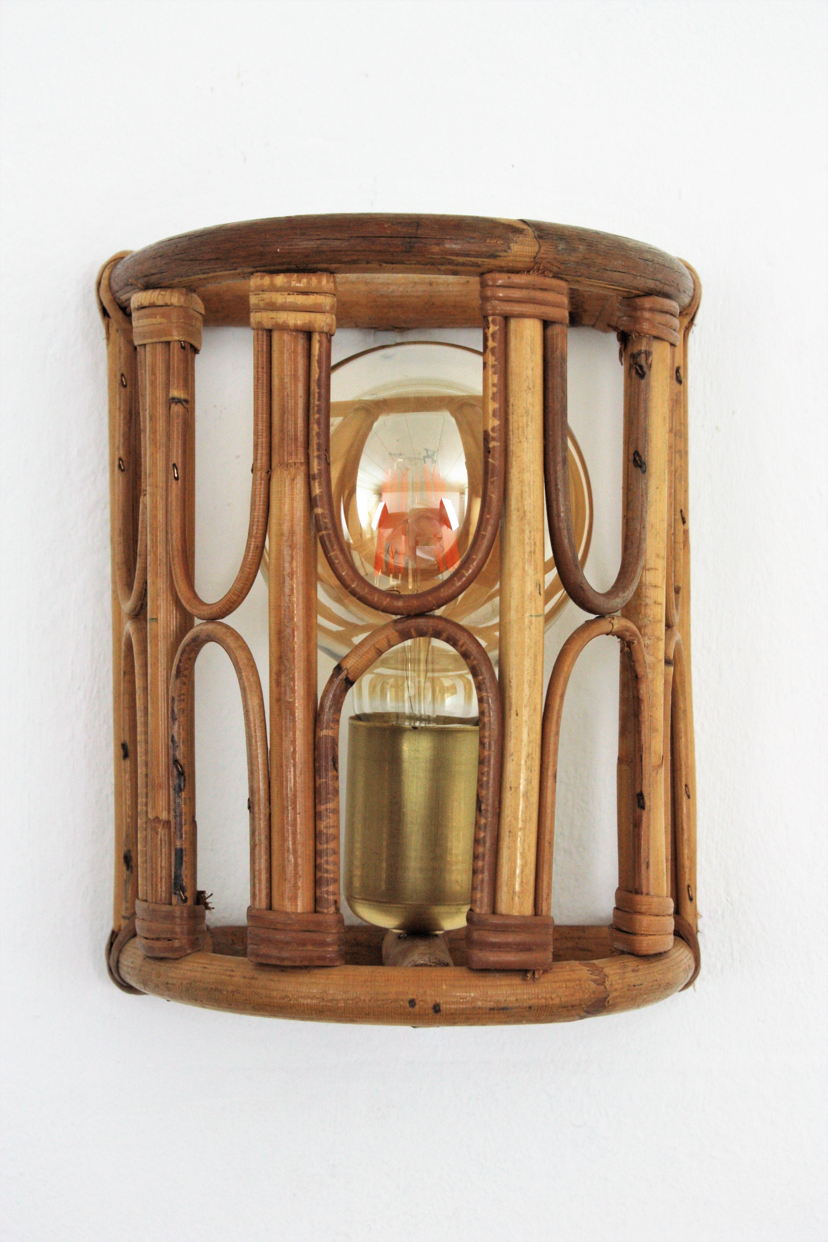 Beautiful half cylinder wall light with bamboo and rattan shades accented by geometric decorations, Italy, 1960s.
This light fixture has a bamboo semi cylindrical structure adorned by rattan details. It has a brass cover to hide the bulb