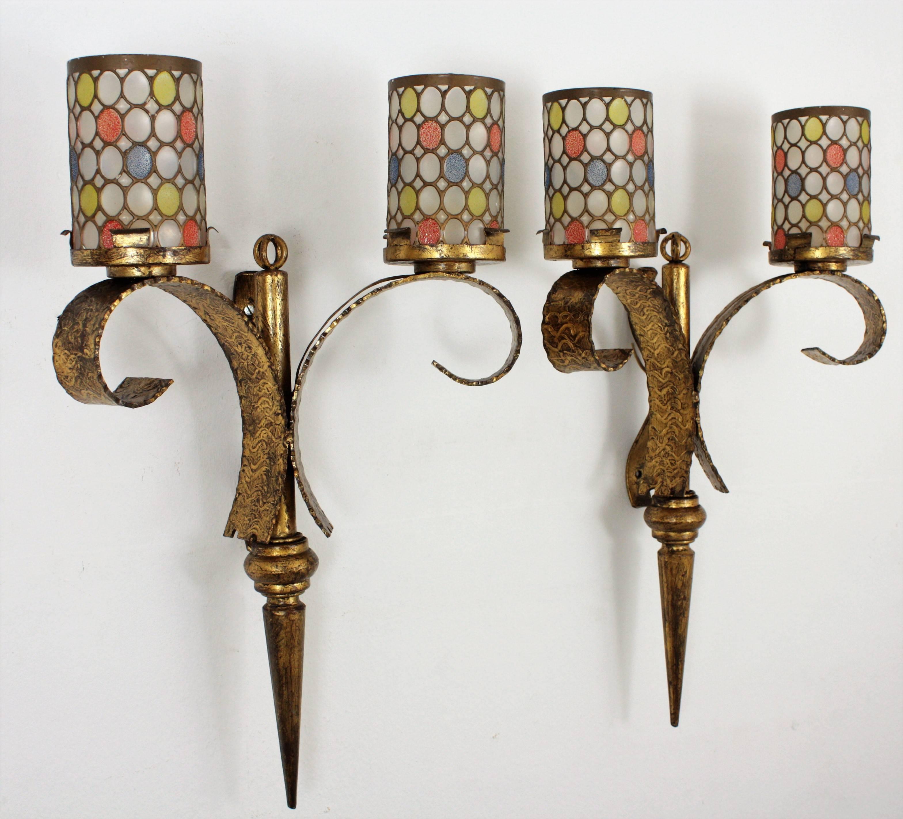 A pair of Hollywood Regency hand-hammered torch shaped gilt iron wall sconces with colorful glass shades with clear glass and yellow, blue and orange accents and two lights, 
Spain, 1940s-1950s.

Dimensions shown are per piece.