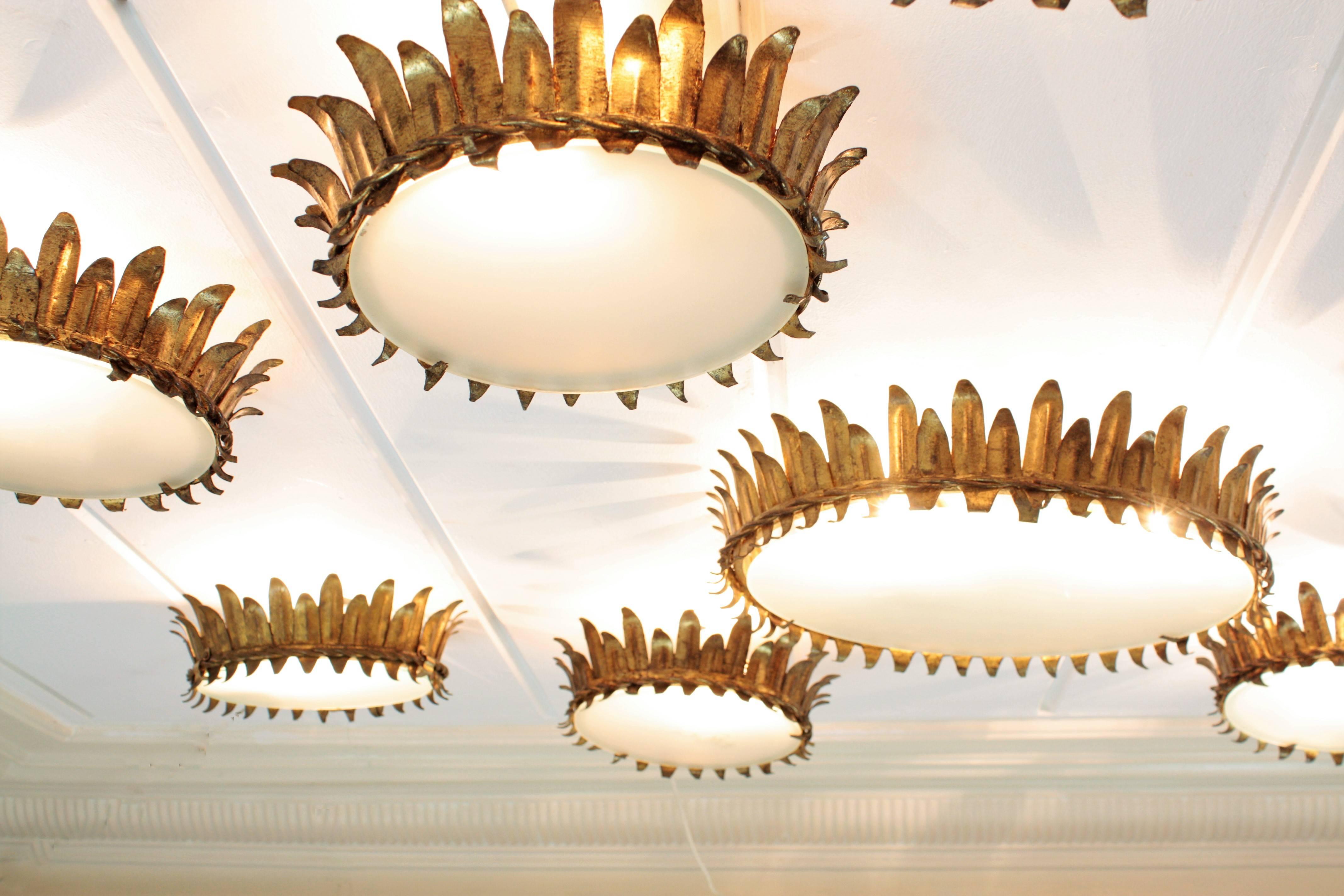 A set of seven magnificent neoclassical gilt iron crown light fixtures or flush mounts. The set is composed by six pieces in the same size and one more in a bigger size. All of them handcrafted in iron and covered with gold leaf. They wear a frosted