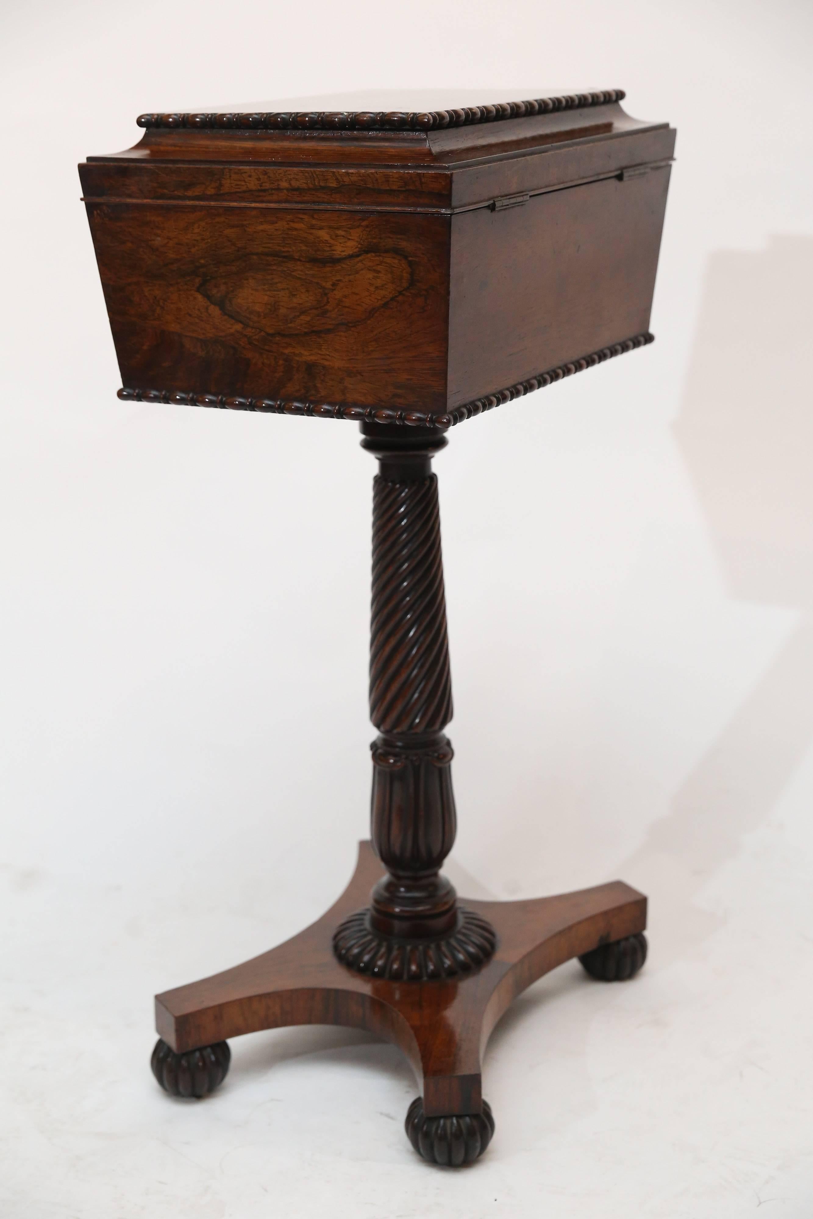 Single pedestal sewing stand is made of rosewood with a beautiful patina.

Box itself is coffin-shaped with a simple carved design around top and bottom perimeter. The top sits on a single fluted leg with four fluted ball feet.

Interior is