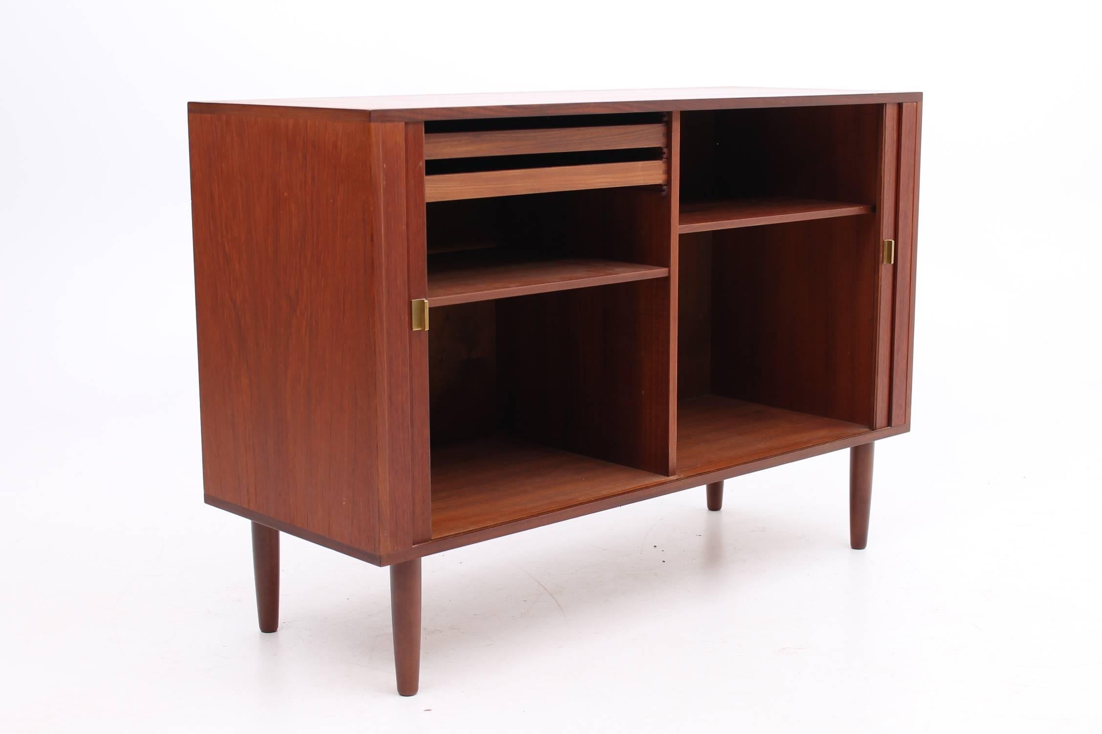 Small sized teak credenza designed by Peter Lovig Nielsen for Hedenstad Furniture. This credenza features brass door pulls to open the roll front drawers. The inside features two organizational slim drawers as well as two fully adjustable shelves.