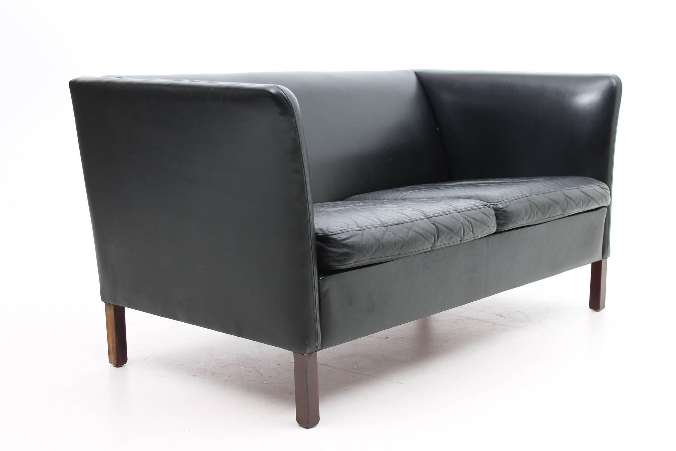 This is a gorgeous black leather loveseat is beautiful from every angle. The black leather is timeless, the curved arms give the loveseat a sofer look, and the wood legs are polished and simple. The loveseat was designed by Hans Olsen for CS Møbler,
