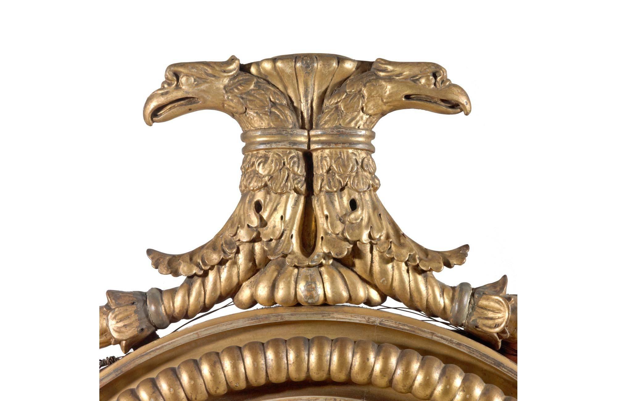 Vergoldeter Konvexspiegel aus dem frühen 19. Jahrhundert. Der innere und äußere Rand mit Verzierungen, das Ganze mit überhöhten Adlerköpfen über Fruchtschnitzereien. In der Mitte der Schürze ein Löwenkopf mit geschnitztem Blumendekor, zwei